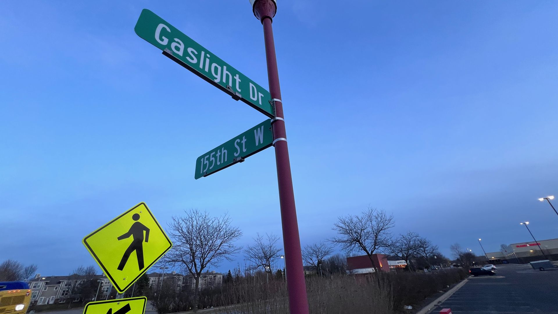 Blue sky over an intersection with green street signs reading Gaslight Dr and 155th St W, a purple street lamp, and bright yellow pedestrian crossing signs; leafless trees line the background.