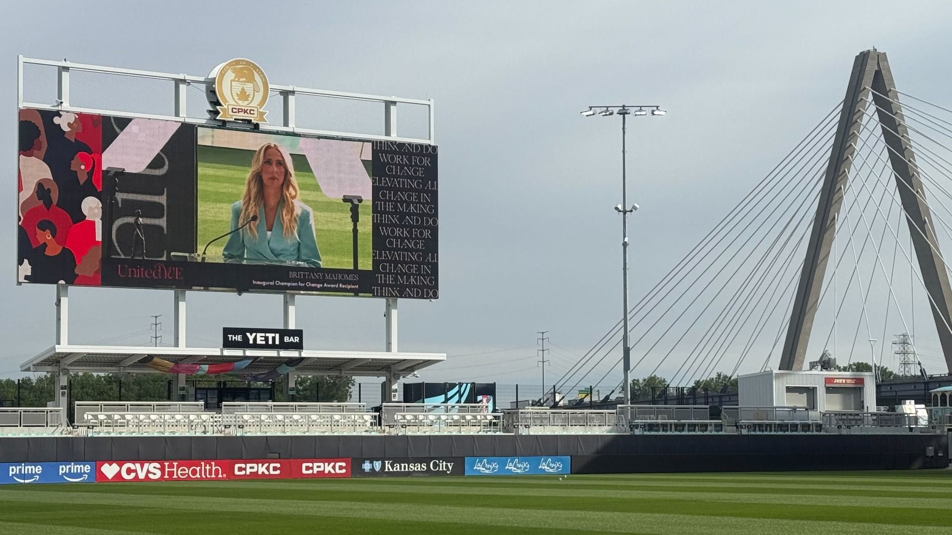 Billboard at a stadium shows a woman in a teal blazer at a podium on a large screen with the words "Think and Do." Left artwork, bottom banners read CVS Health and CP KC; a cable-stayed bridge stands on the right.