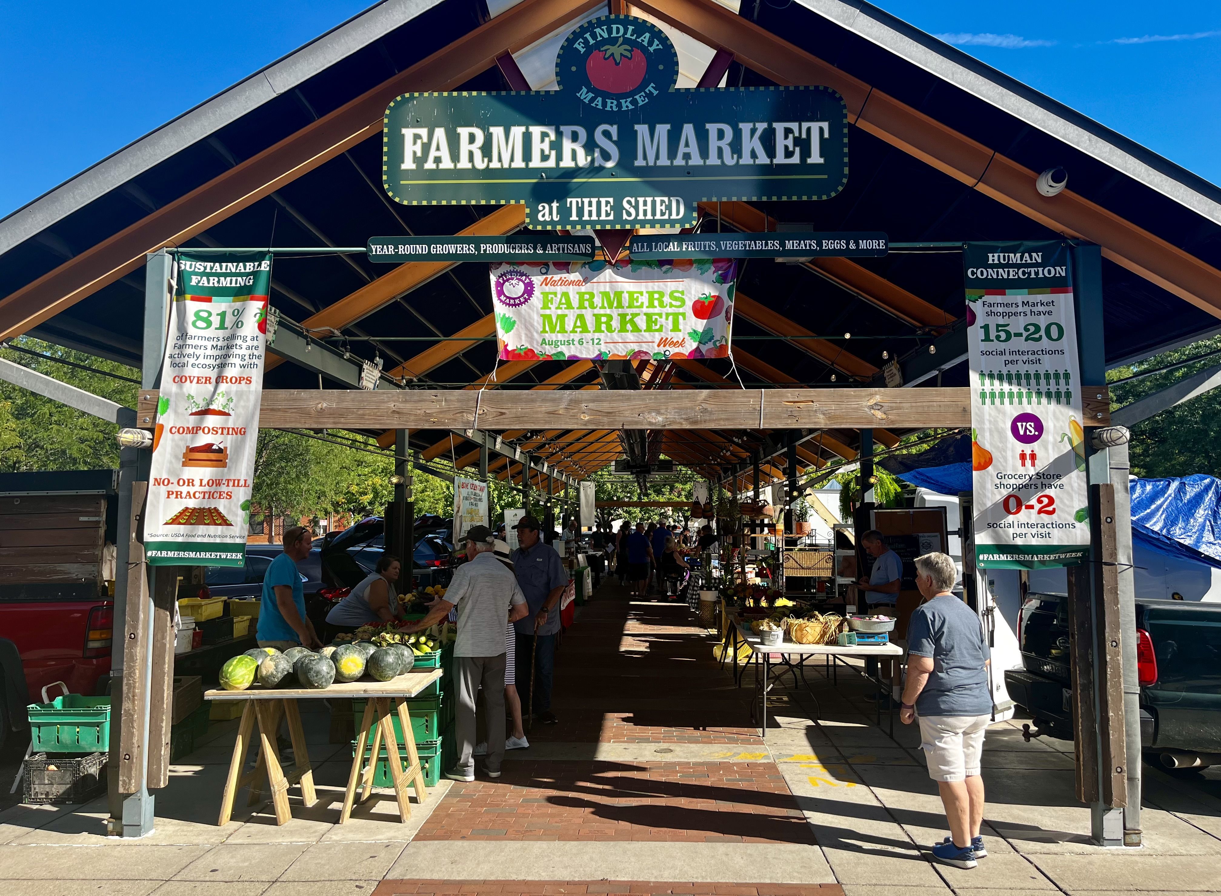 Open-air farmers market under a wooden pavilion with a large sign reading "FARMERS MARKET at THE SHED." Colorful banners promote sustainable farming as shoppers browse fruits and vegetables.