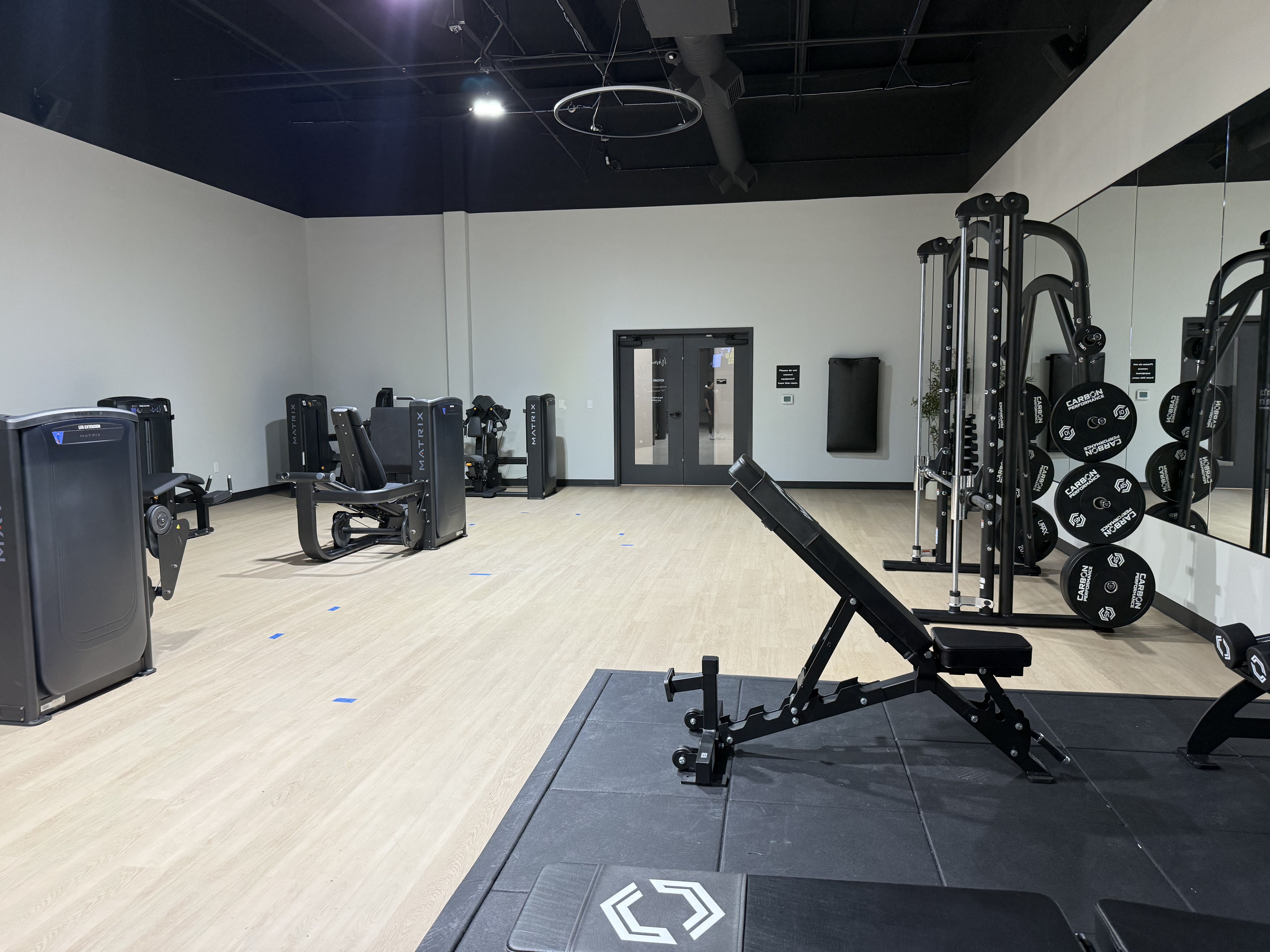 Gym interior with light wood floor, gray walls with a dark blue upper trim, large right-side mirror, and black weight machines with plates; ceiling shows ducts and a circular light.