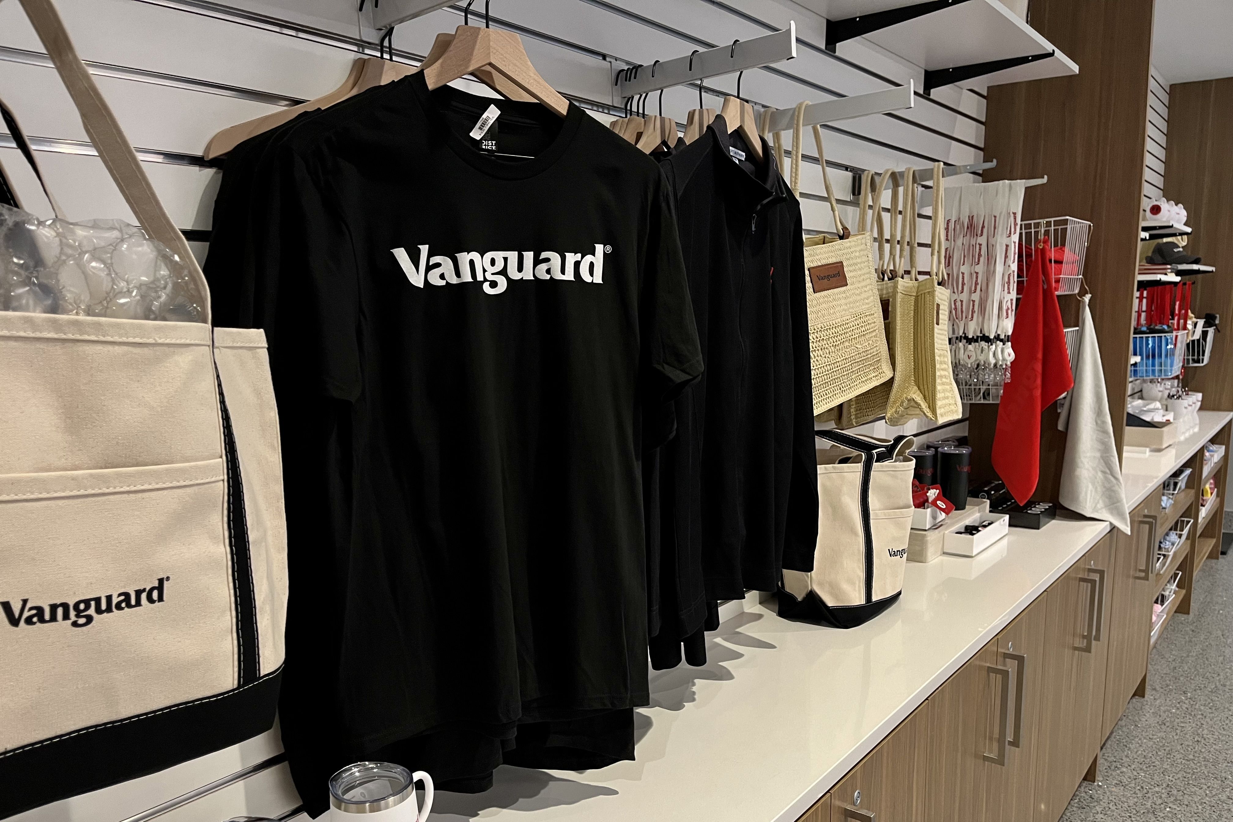 Shop display with black "Vanguard" t-shirts, beige totes, woven bags, and various accessories on white counter and wood shelves.