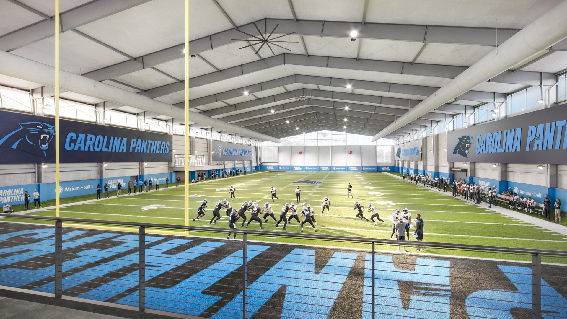 Indoor football field for the Carolina Panthers; players practice on green turf, with blue banners reading CAROLINA PANTHERS along the walls, a blue track around the field, and large ceiling fans.