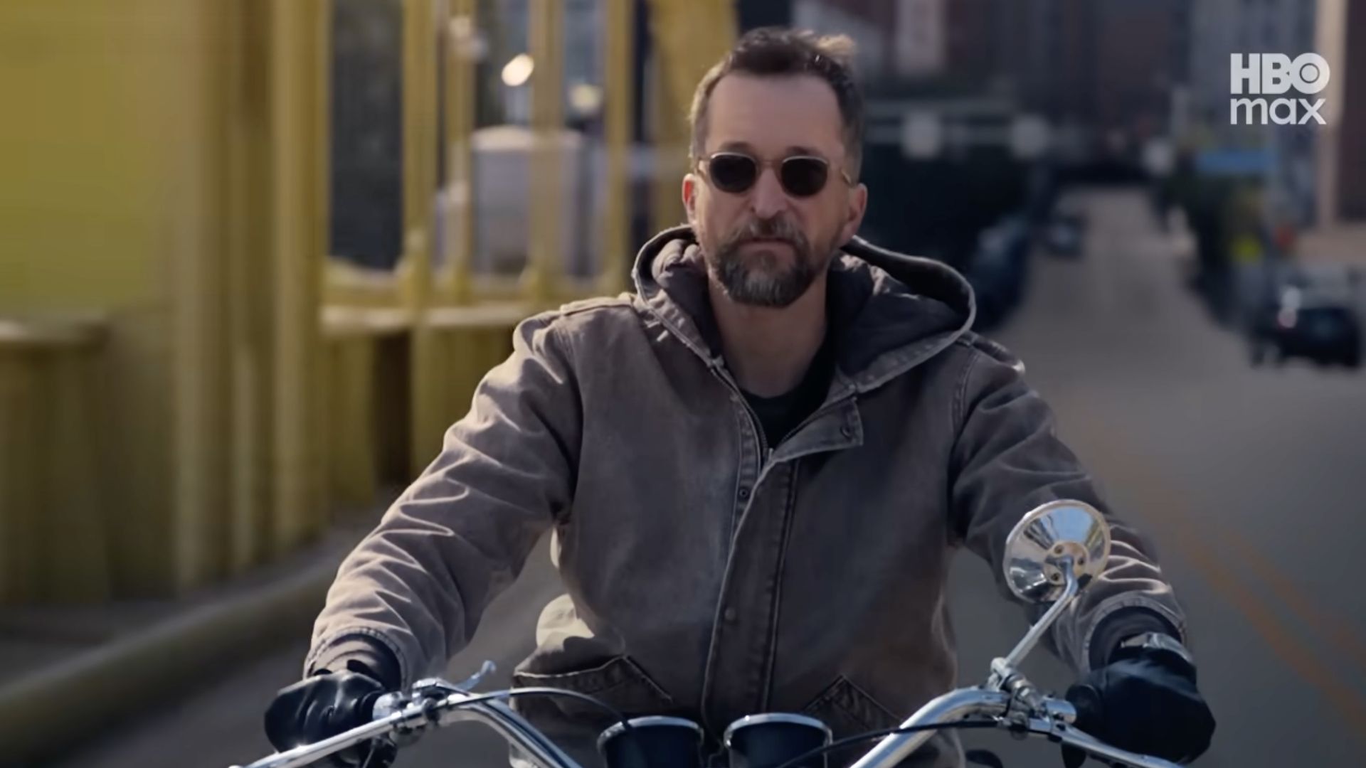 Man wearing sunglasses, brown jacket, and black gloves rides a motorcycle on a street with yellow poles and an HBO Max logo in the upper right corner.