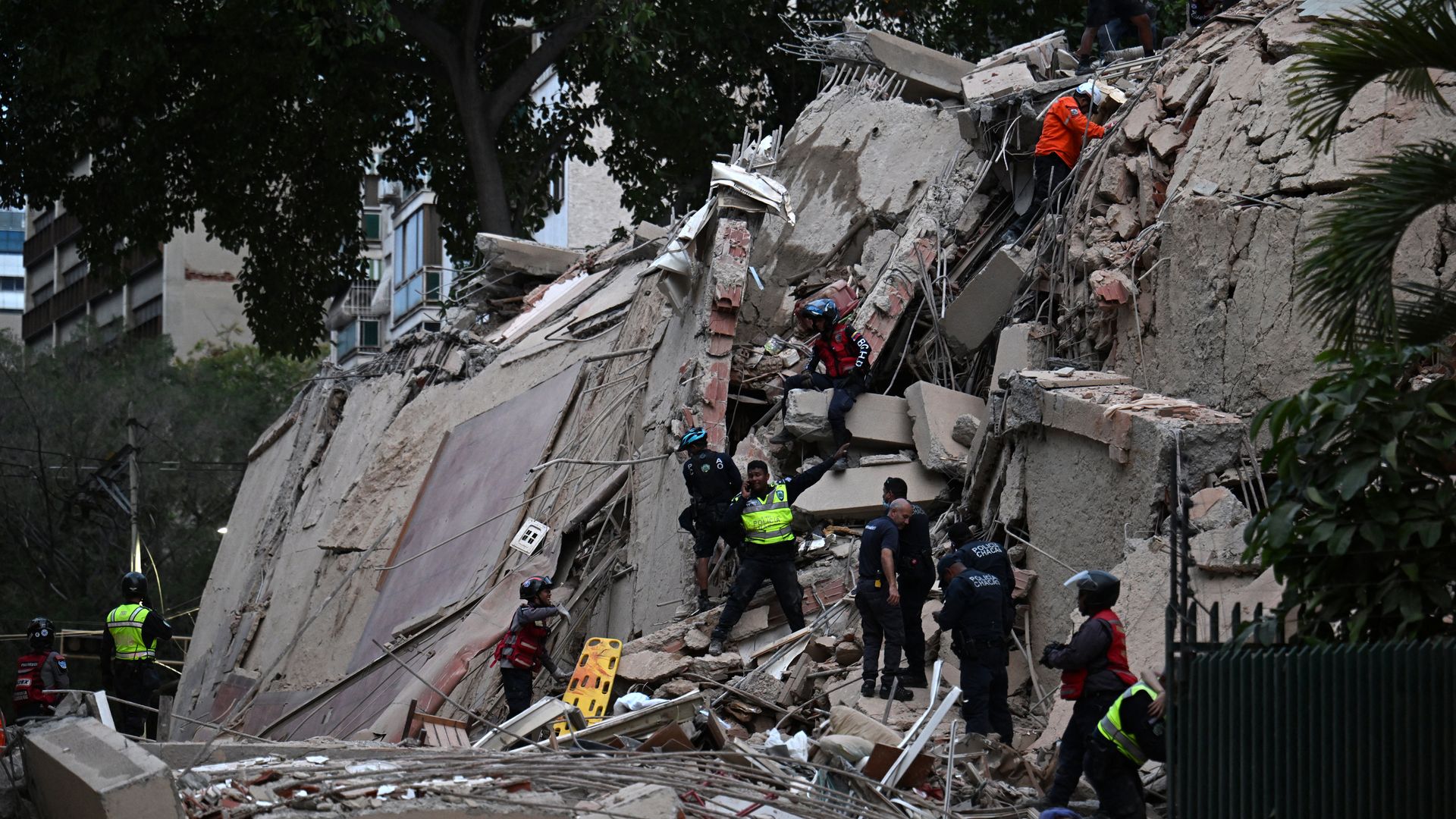 Rubble from a collapsed building on a slope with rescue workers in orange vests and helmets and police scanning the debris; trees and urban buildings visible in the background.