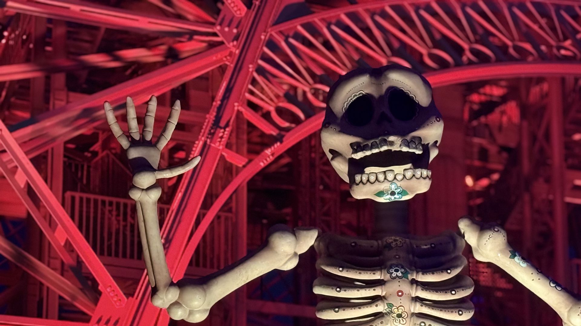 Life-size skeleton with a sugar-skull face and floral patterns on its bones raises a hand, set against a red-lit framework of metal beams and scaffolding.