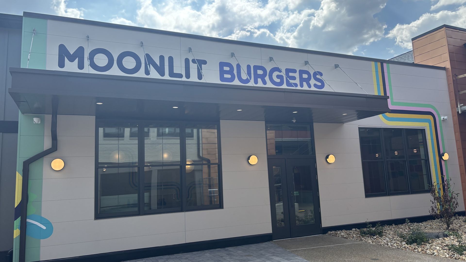 Exterior of Moonlit Burgers restaurant with white facade, blue sign, circular wall lights, and colorful striped design on right side under partly cloudy sky.