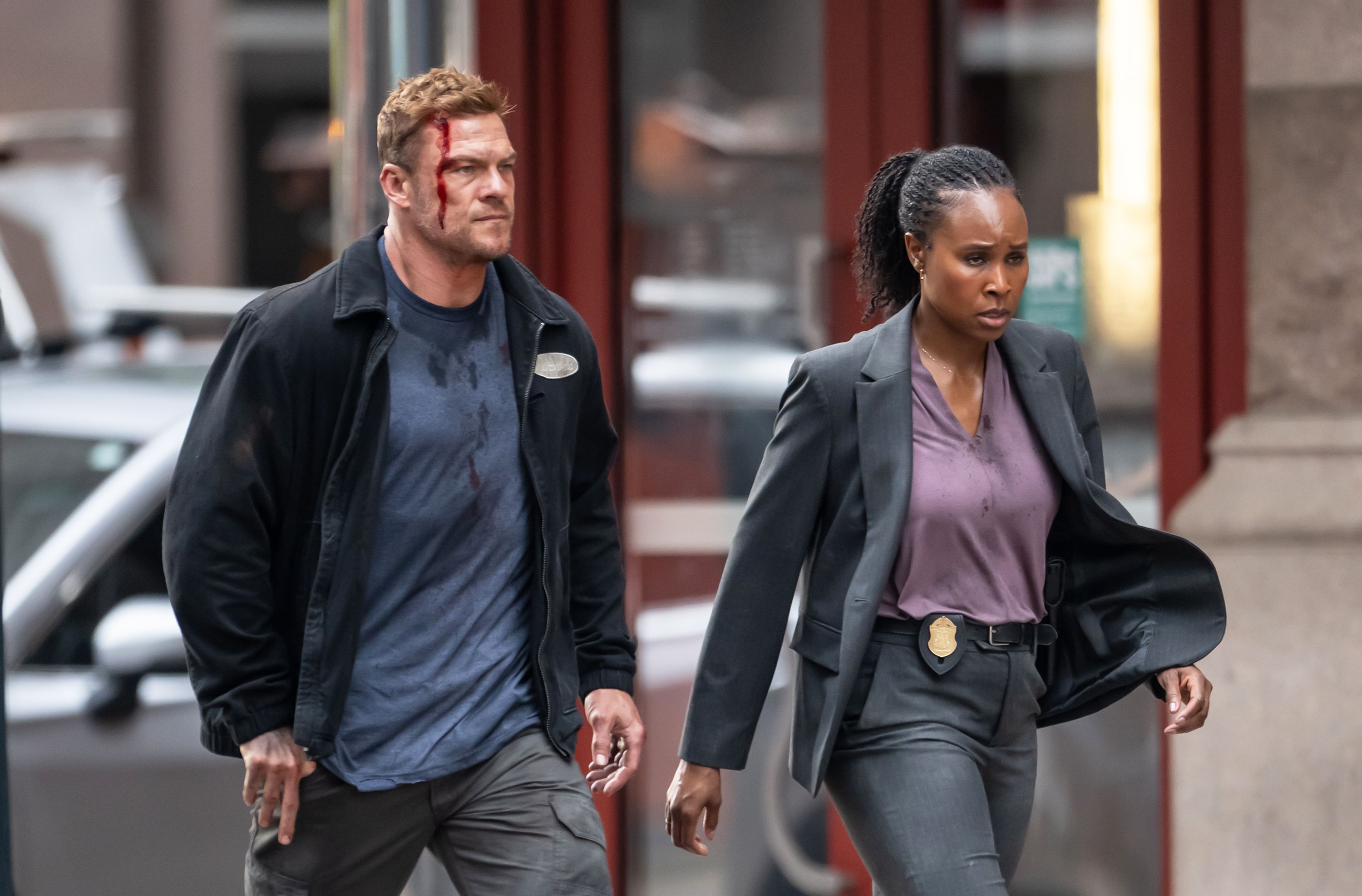 Two serious-looking people walking on a city street; man with blood on his face and shirt wears casual clothes, woman in grey suit has a police badge on her belt.