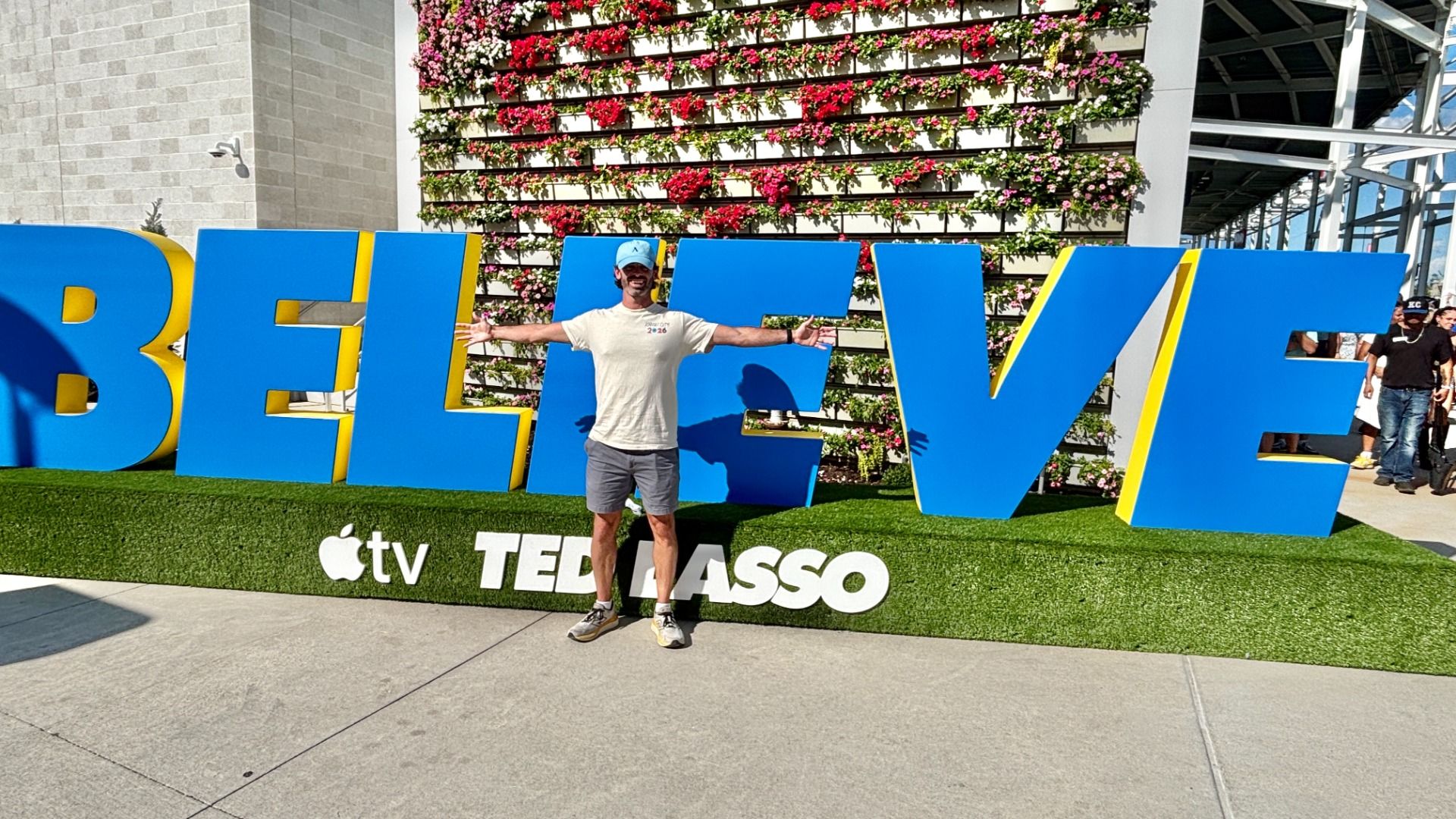 Man in a gray T-shirt and blue cap stands with arms out in front of giant blue "BELIEVE" letters with yellow edges at an Apple TV TED LASSO event, against a flower wall.