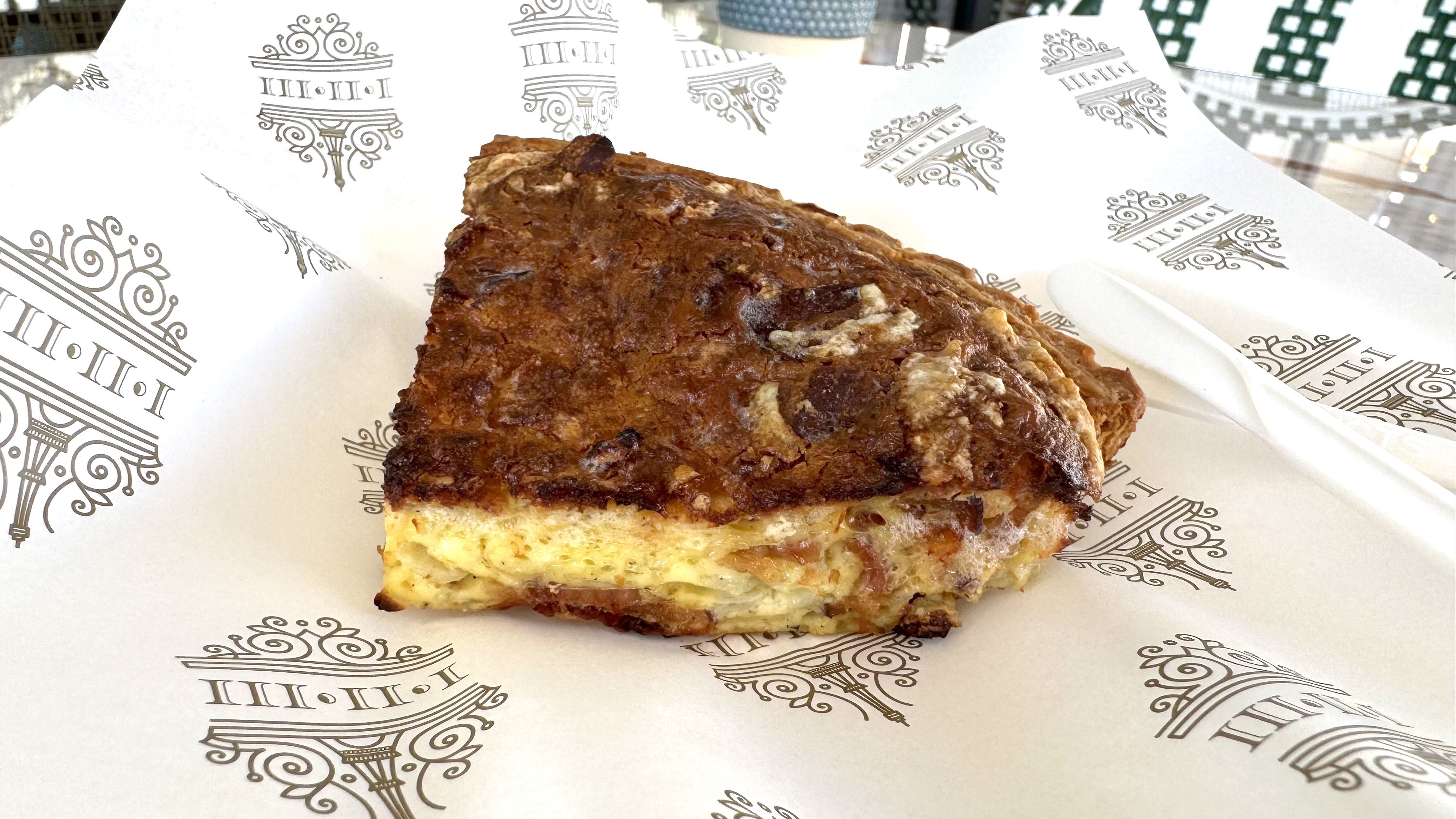 A slice of golden-brown quiche with a crispy top and fluffy interior sits on decorative parchment paper next to a plastic knife.