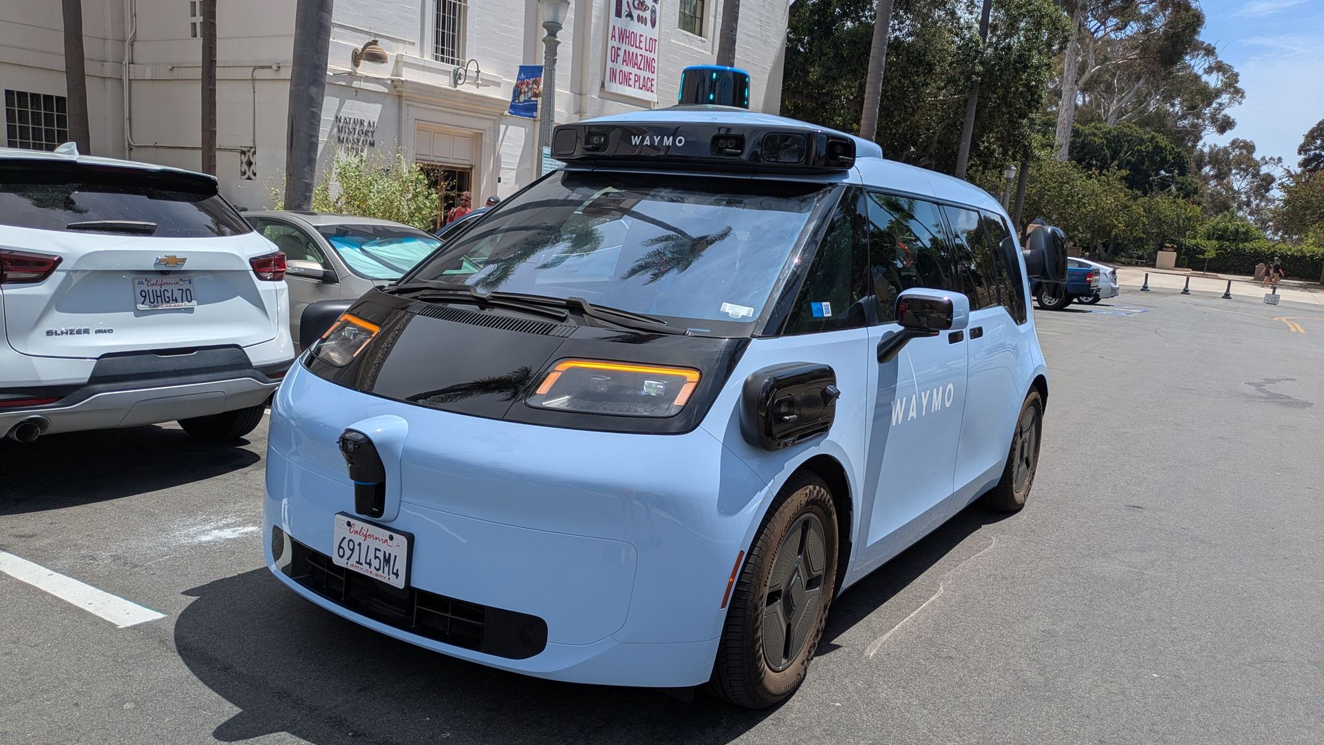 Blue Waymo autonomous van with a blue top light parked in a lot beside a white museum building, flanked by palm trees and other cars.