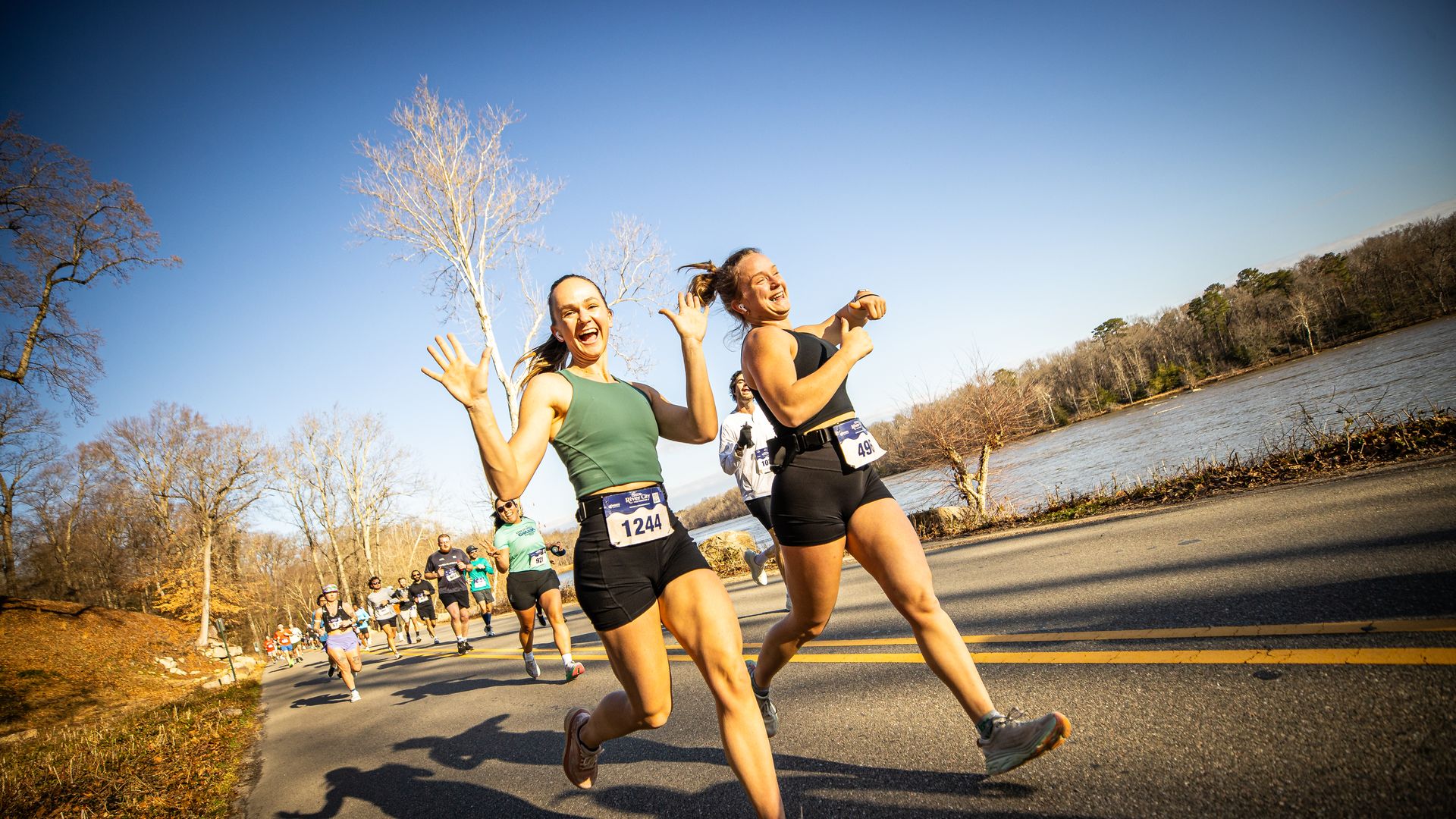 Two women joyfully running a road race near a river under a clear blue sky, wearing race bibs 1244 and 491 with other runners behind them on a paved road lined with leafless trees.