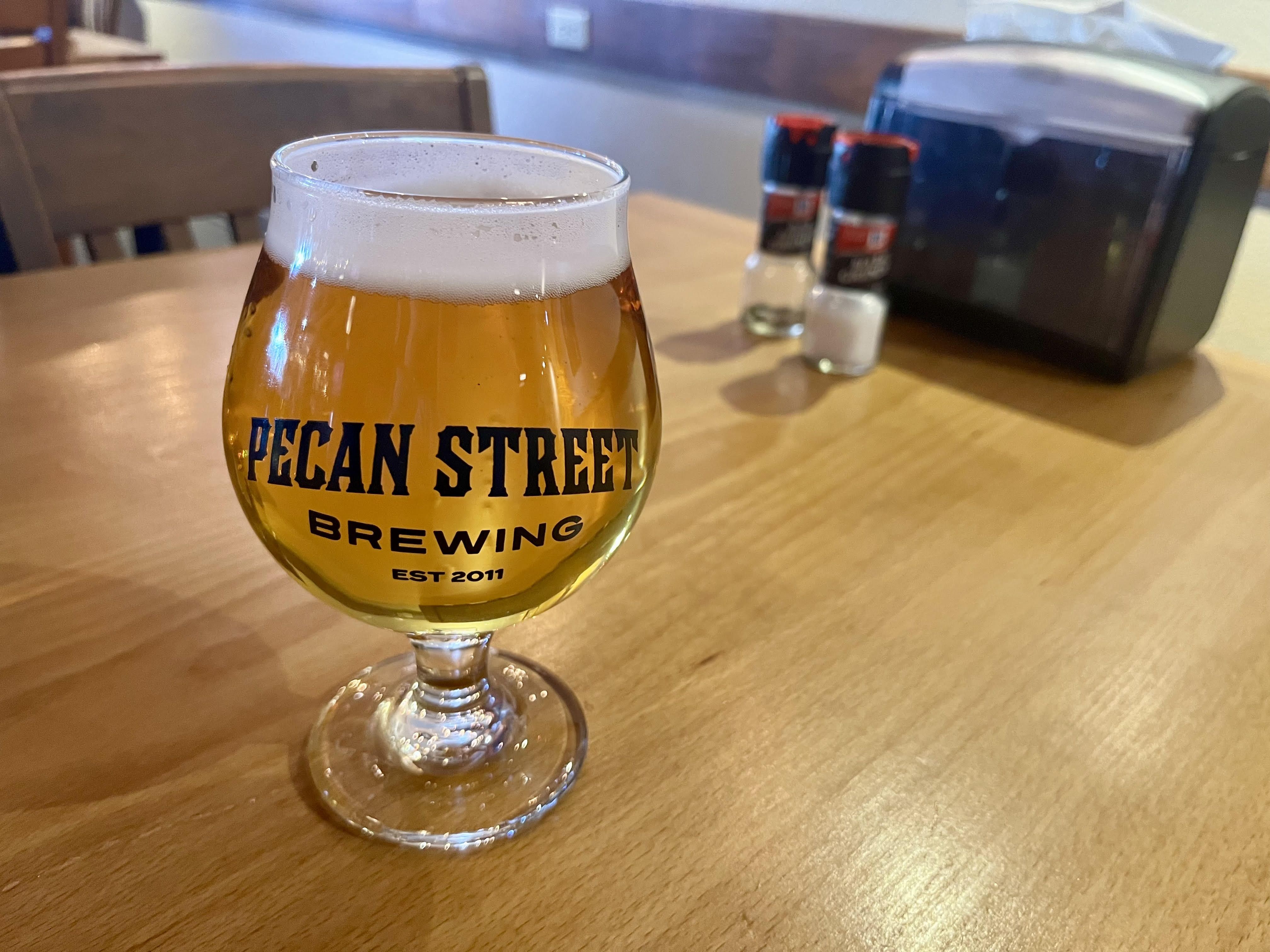 A beer glass holds a light colored beer with foam. It reads Pecan Street Brewing.