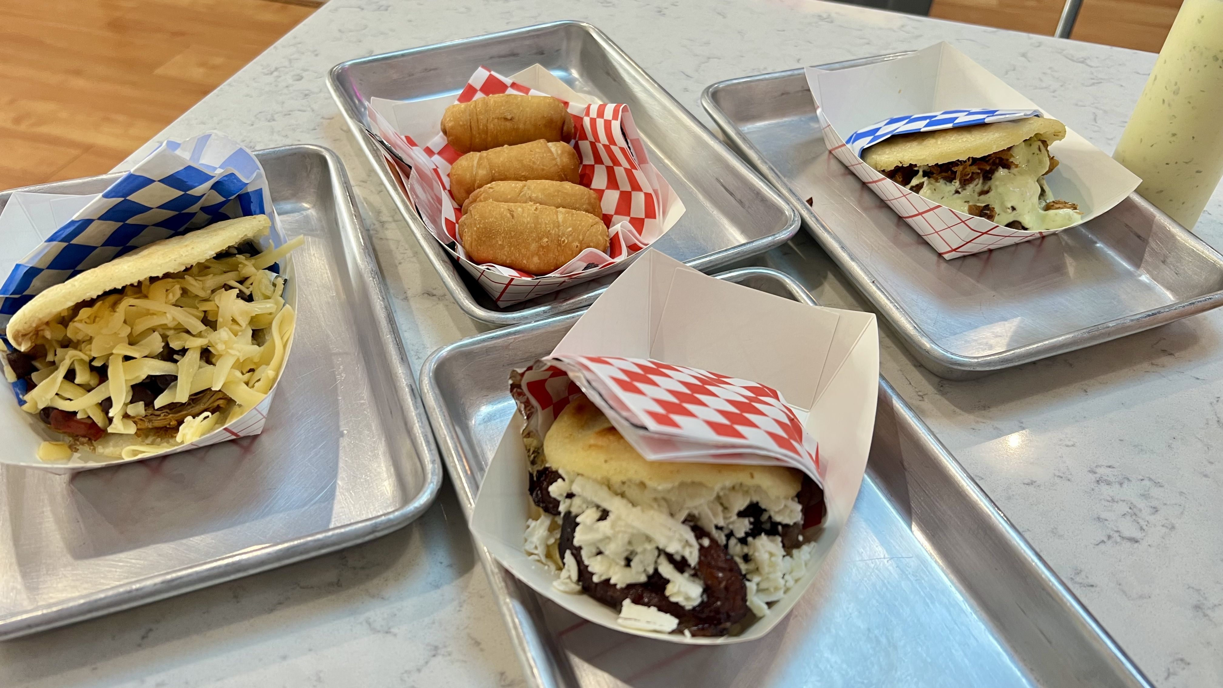 Three arepas with different fillings and cheese on metal trays, along with a tray of four fried finger foods, all on a light-colored countertop.