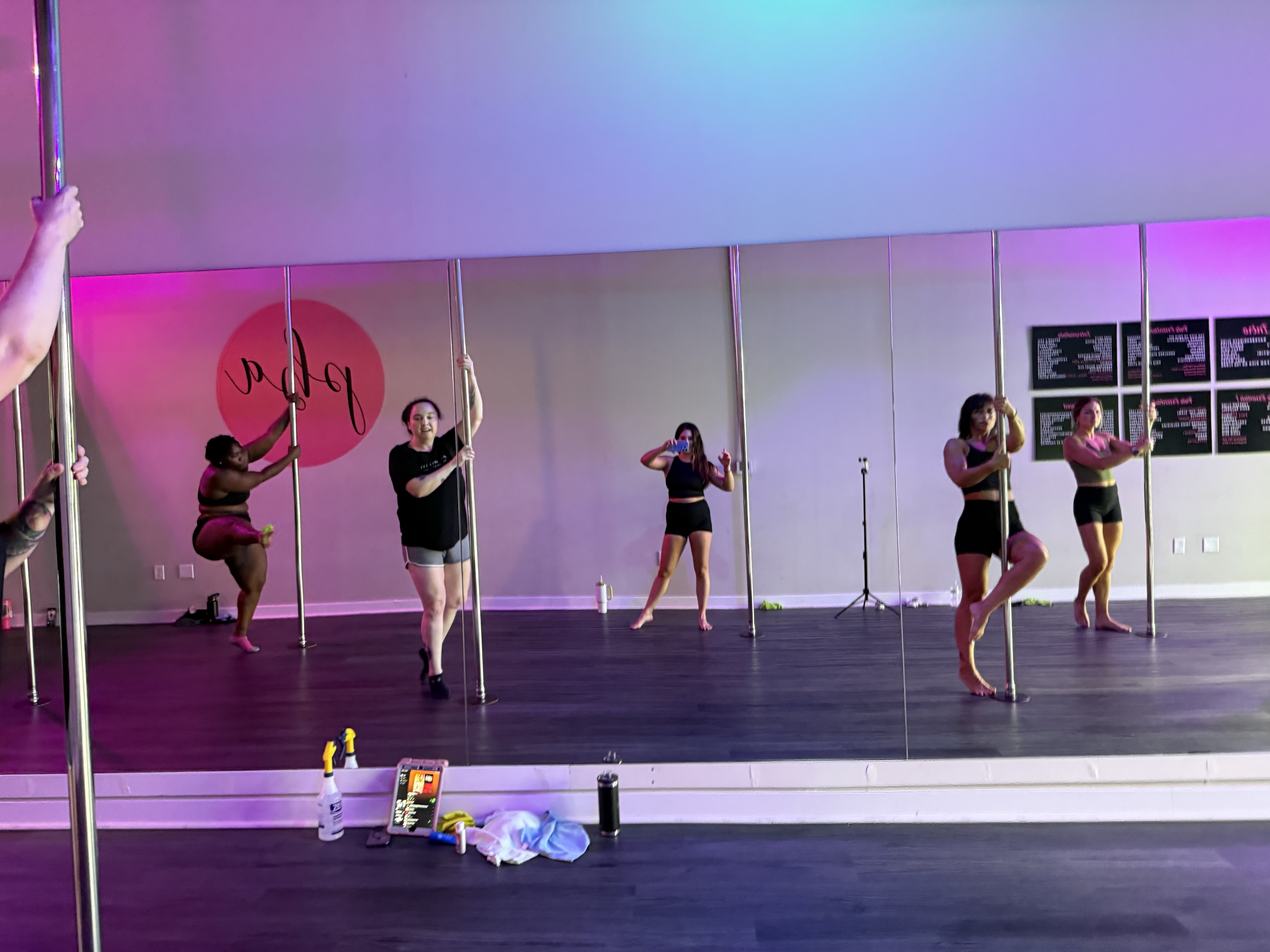 Group of women in a pole dancing studio holding poles, with purple and blue lighting, mirrored wall, and various personal items on the floor in the foreground.