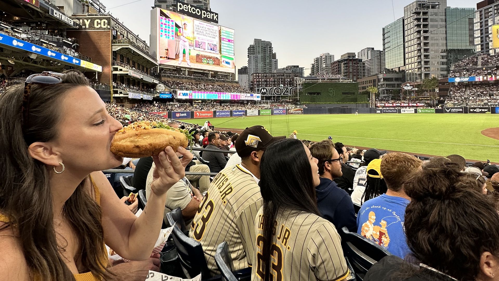 A woman sitting in the stands at a baseball game eats a hotdog.