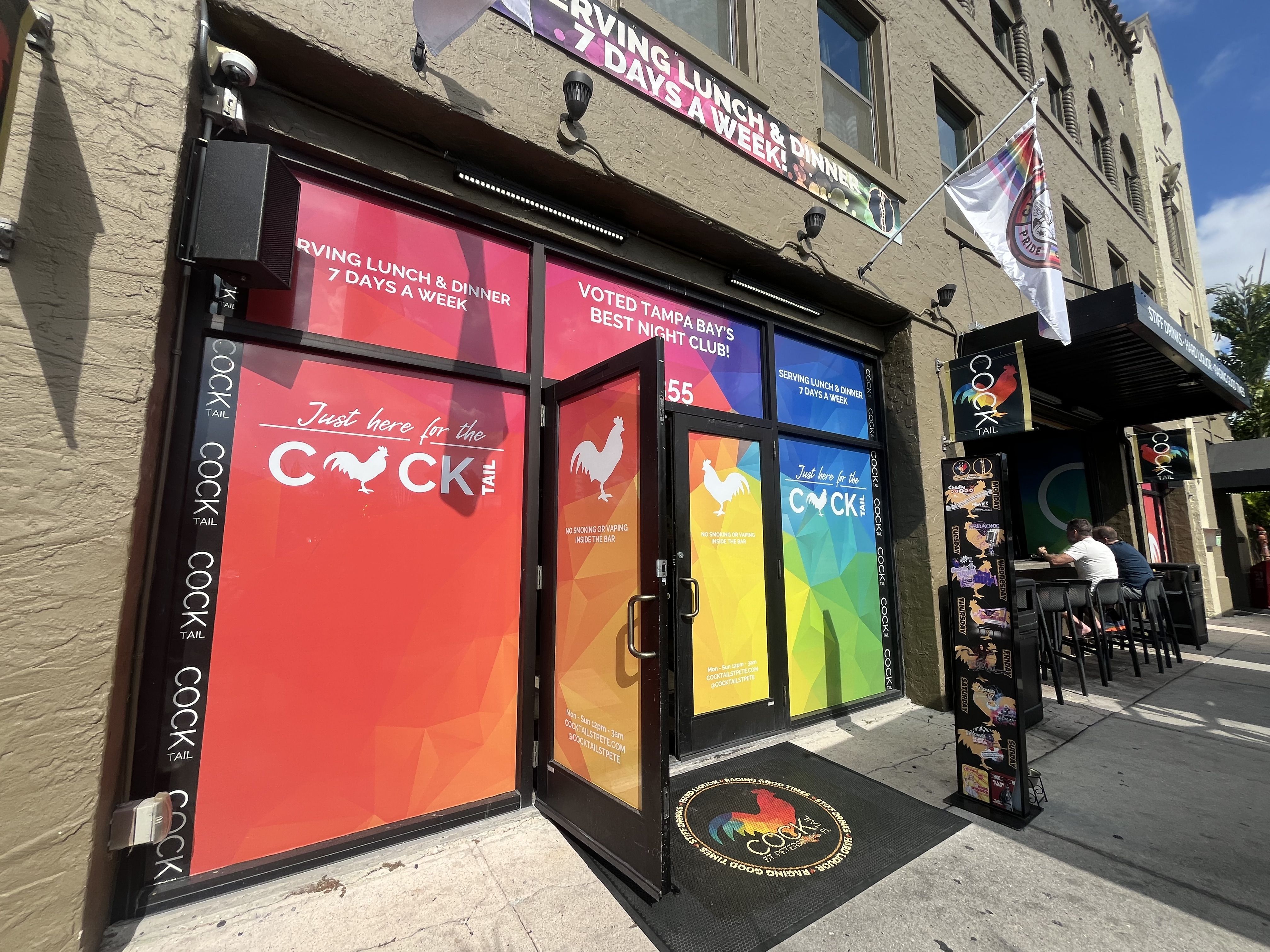 Front entrance of "COCKTAIL" nightclub with rainbow-colored windows, a rooster logo, text about serving lunch and dinner 7 days a week, and patrons sitting outside on chairs.