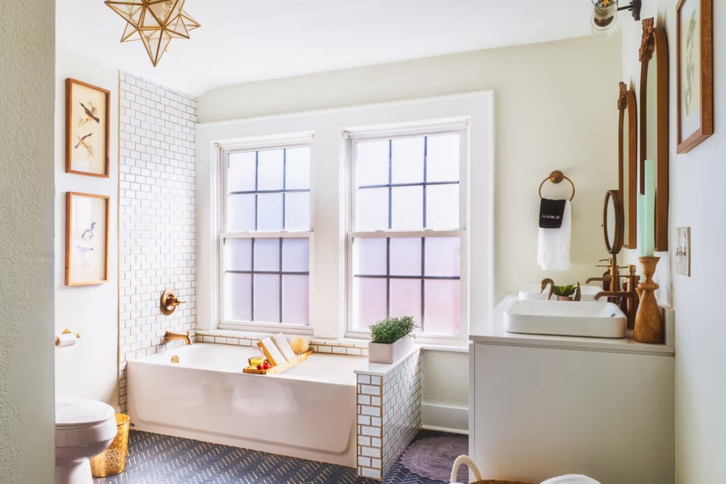 Bright bathroom with white subway tiles, gold fixtures, bathtub under two large windows, plant on tub, dual sinks with wooden-framed mirrors, bird artwork, and star-shaped pendant light.