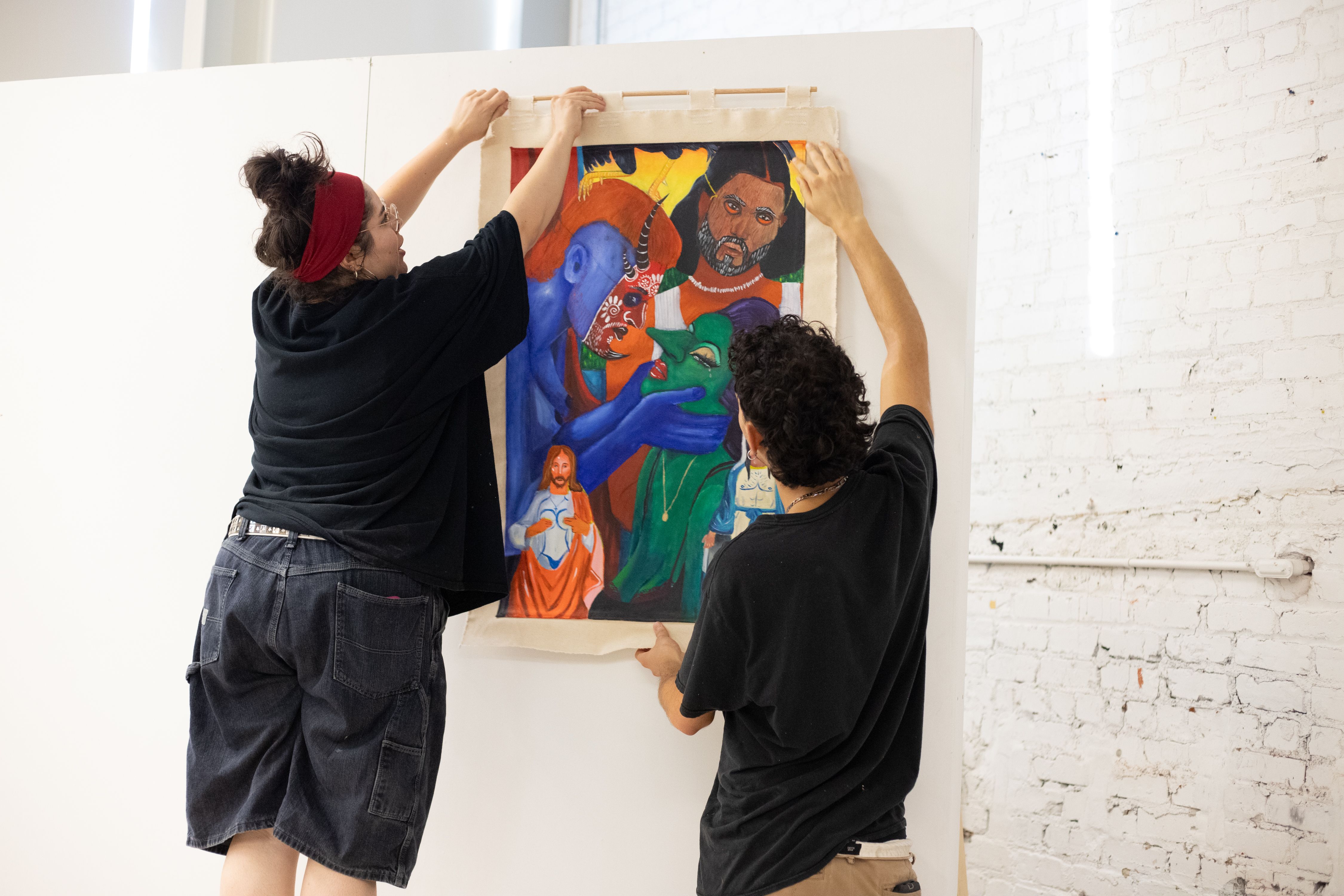 Two people hanging a colorful painting on a white wall, depicting blue, green, orange, and brown faces with detailed expressions.