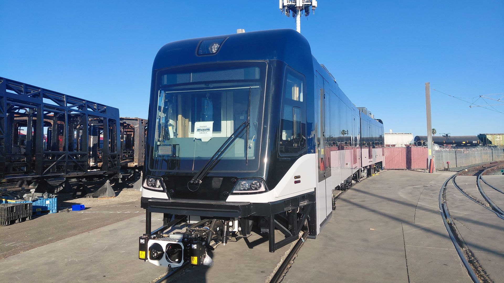 Blue electric train seen from the front in a rail yard, with tracks, a tall white antenna tower behind it, and a clear blue sky.