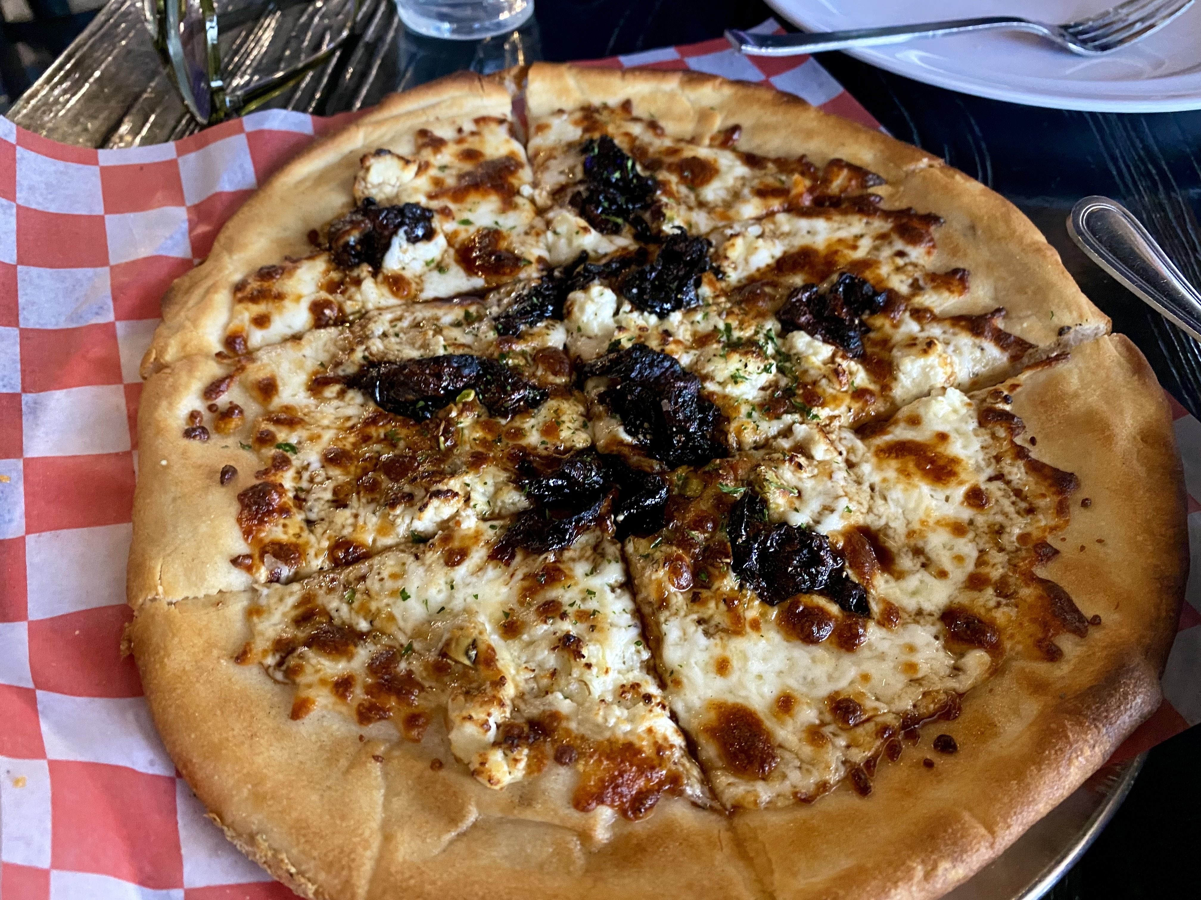 A pizza with a golden crust topped with bubbly, melted cheese and scattered pieces of dark, sun-dried tomatoes, placed on red and white checkered paper on a metal tray.