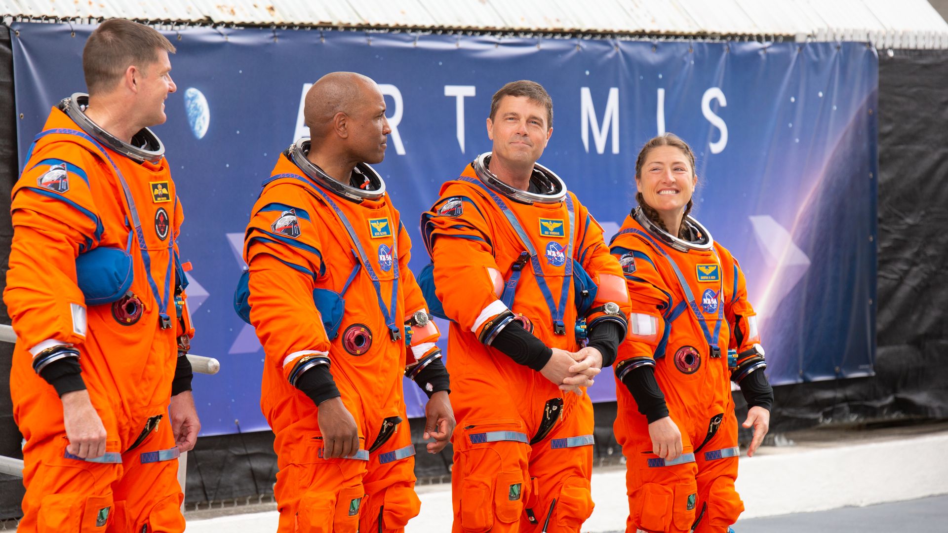 Four astronauts in bright orange spacesuits stand in a row outdoors, helmets tucked around their necks, patches visible on suits, with a blue Artemis banner in the background.