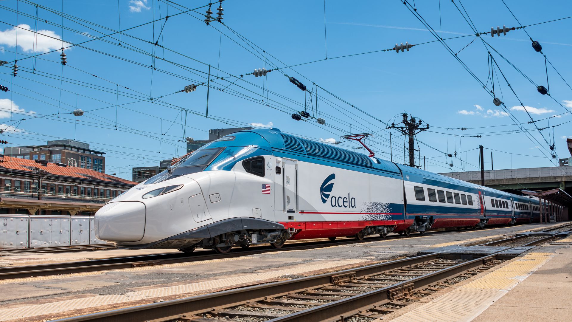 A sleek Acela high-speed train with white, blue, and red colors parked at a train station under a bright blue sky with overhead wires and surrounding buildings.