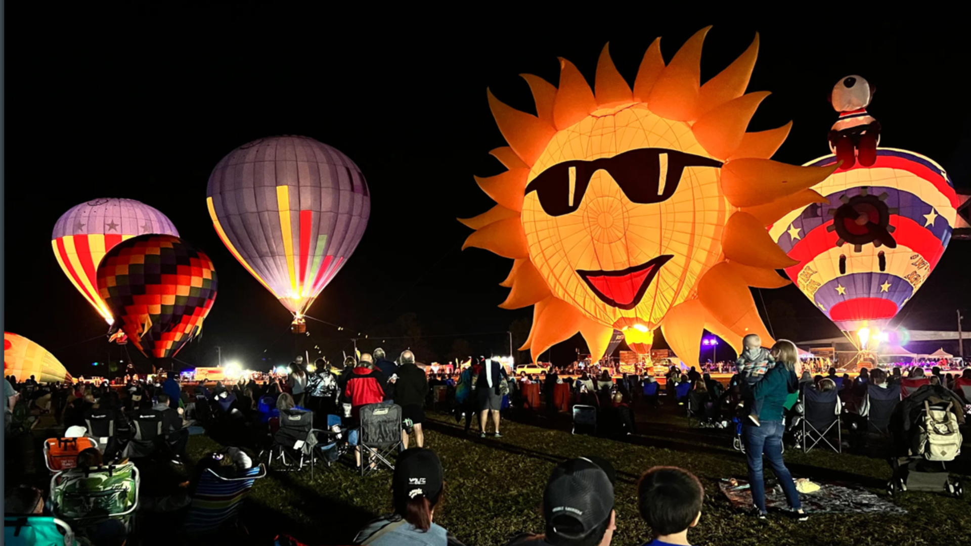 Image shows a night-time picture of a sun hot air balloon and others filled with air.