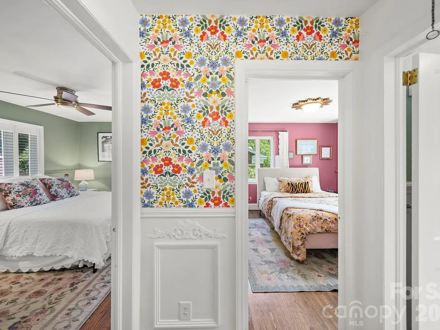 View down a hallway showing two bedrooms through doorways; a central wall is covered in bright floral wallpaper. Left room: green walls, white bedding. Right room: pink walls, floral quilt.
