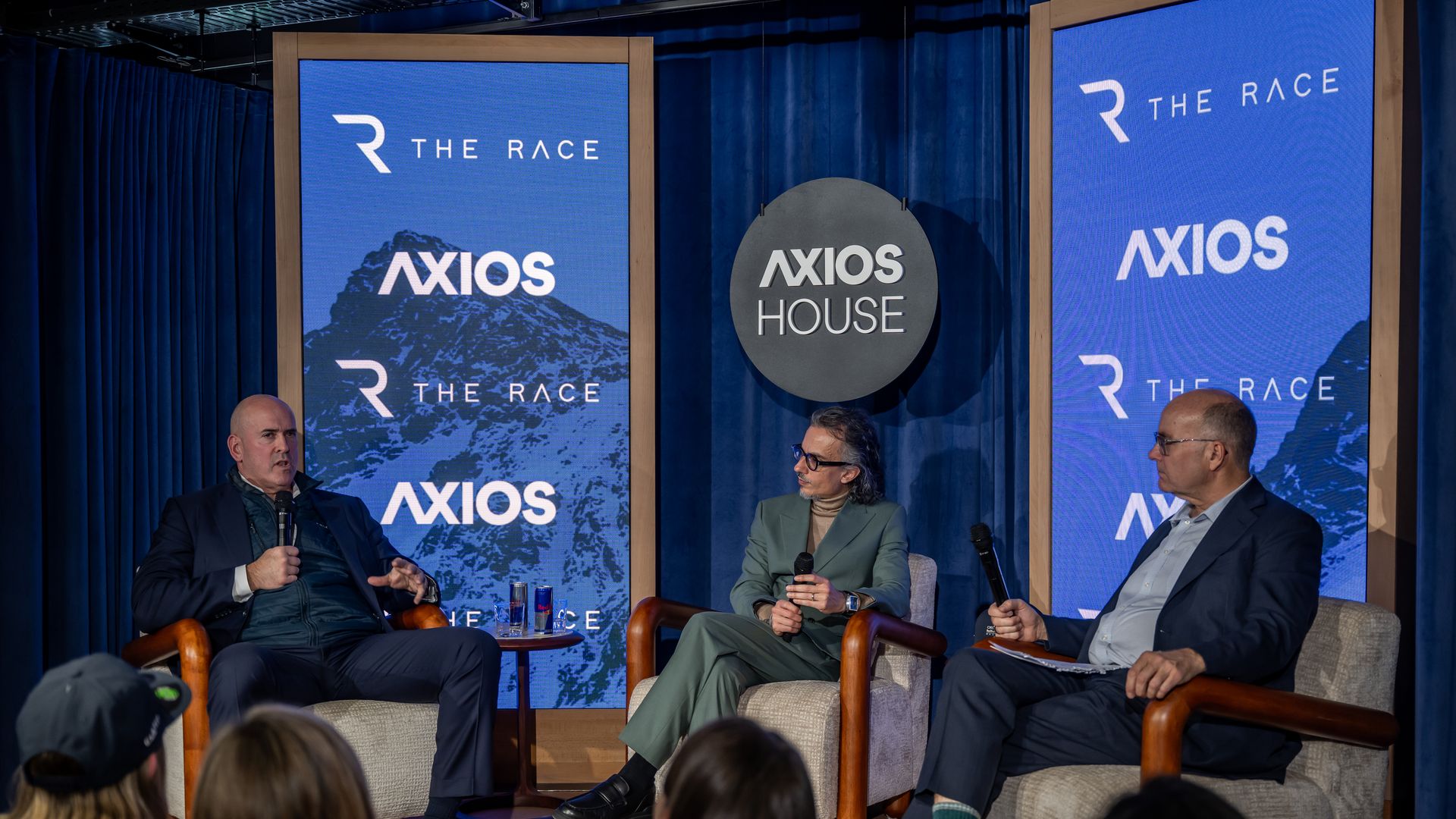 Three men holding microphones sit on armchairs on a stage with blue curtains and screens displaying "THE RACE" and "AXIOS" logos. Audience heads are visible in foreground.