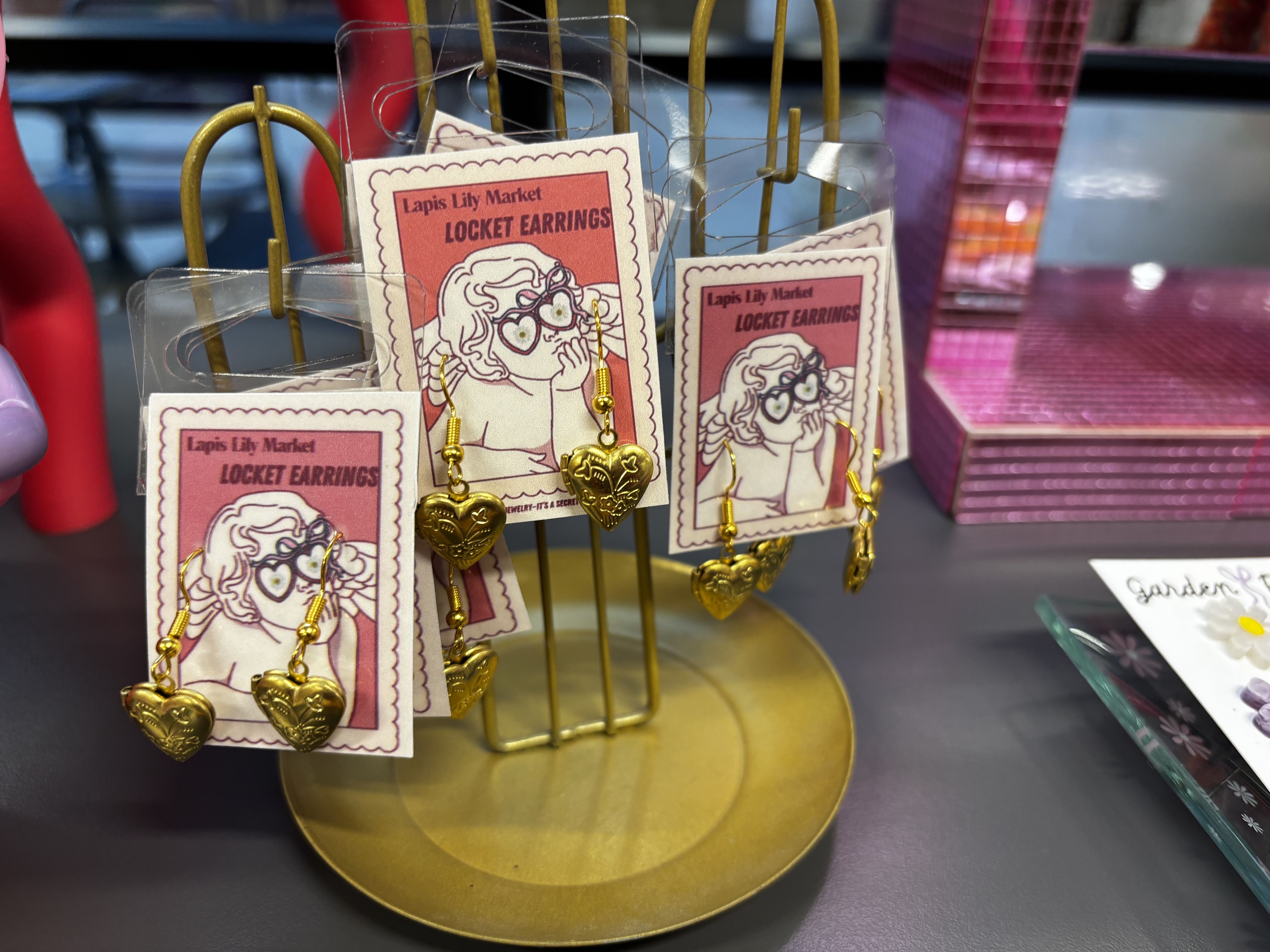 Gold heart-shaped locket earrings hanging on a yellow metal stand, with pink cards labeled "Lapis Lily Market LOCKET EARRINGS" featuring a sketch of a person wearing heart-shaped glasses.