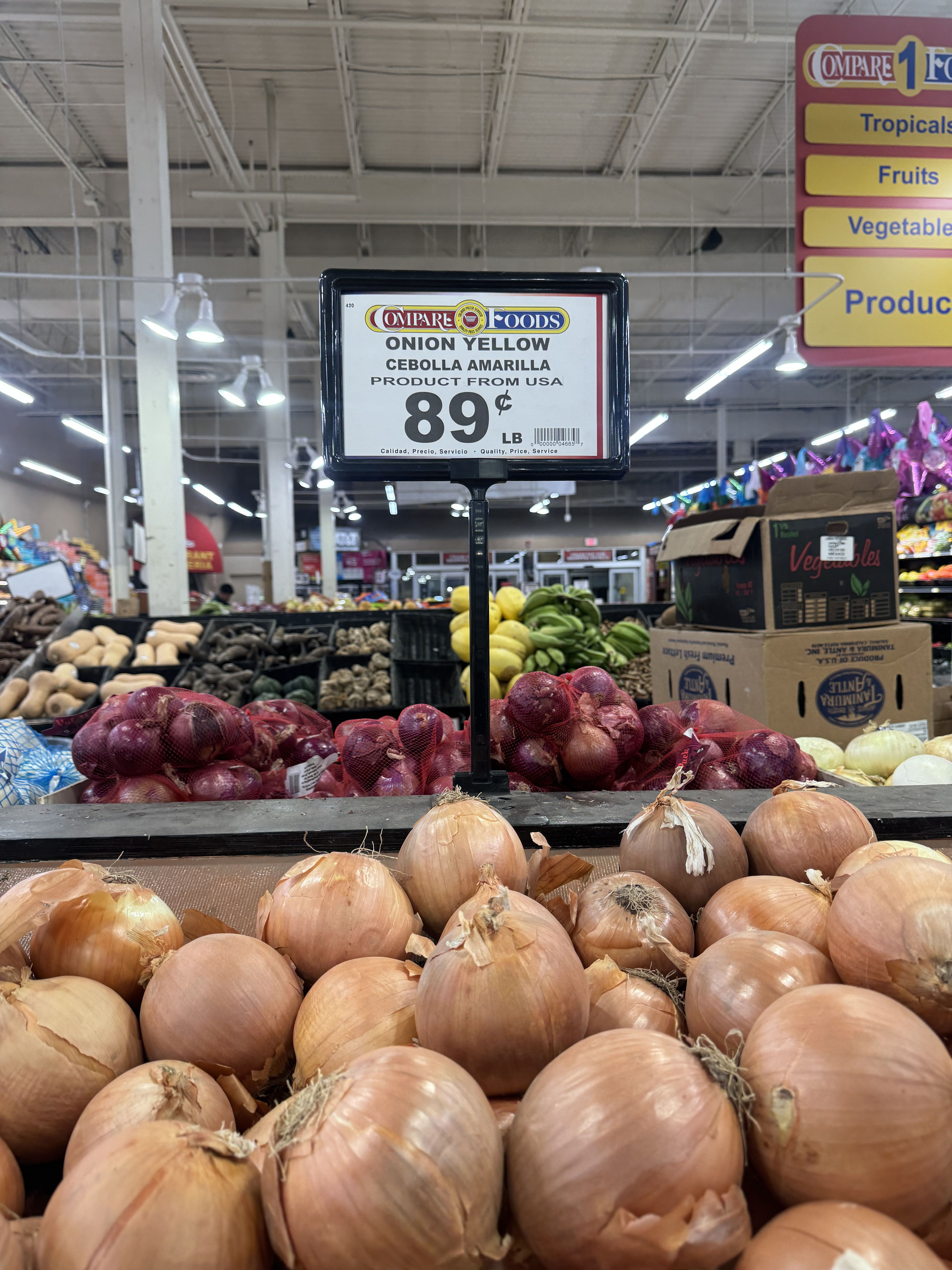Yellow onions displayed in a supermarket with a sign showing price 89 cents per pound, labeled in English and Spanish, with other produce and colorful signs visible in the background.
