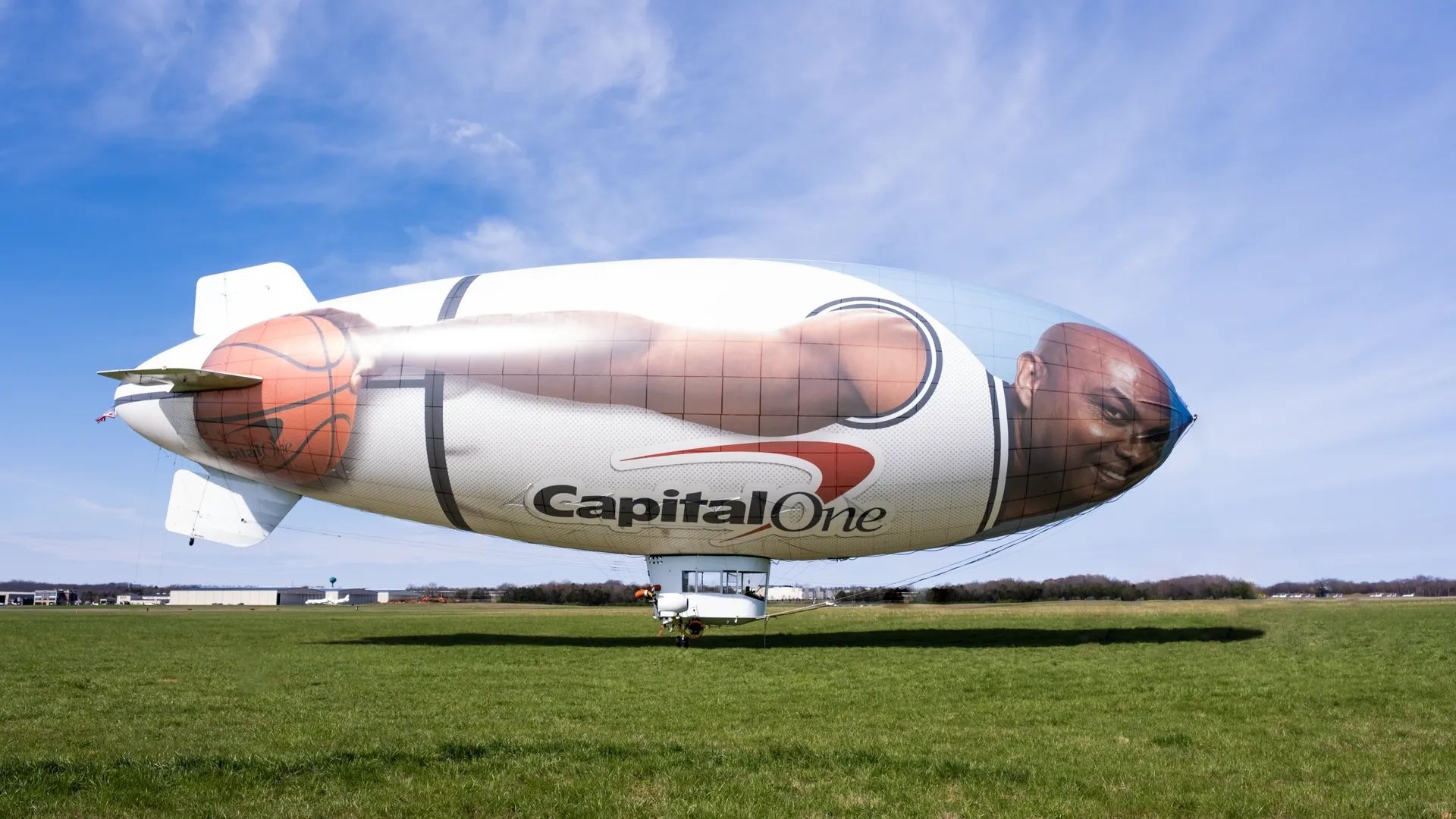 A large white airship sits on a grassy field under a blue sky, featuring a photo of Charles Barkley, with the Capital One logo along its side.
