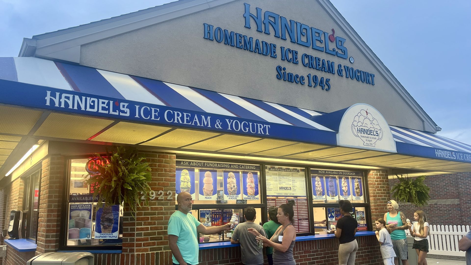 Exterior of Handel's Homemade Ice Cream & Yogurt shop with a blue sign, striped blue-and-white awning, brick walls, and customers lined up at the counter under a bright sky.