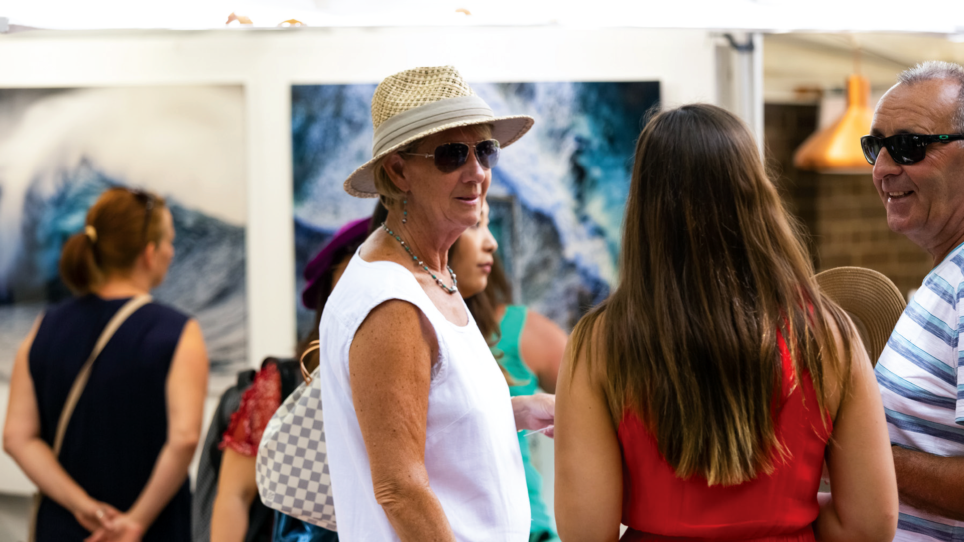 A group of people talks in an art gallery, with paintings of surf displayed in the background
