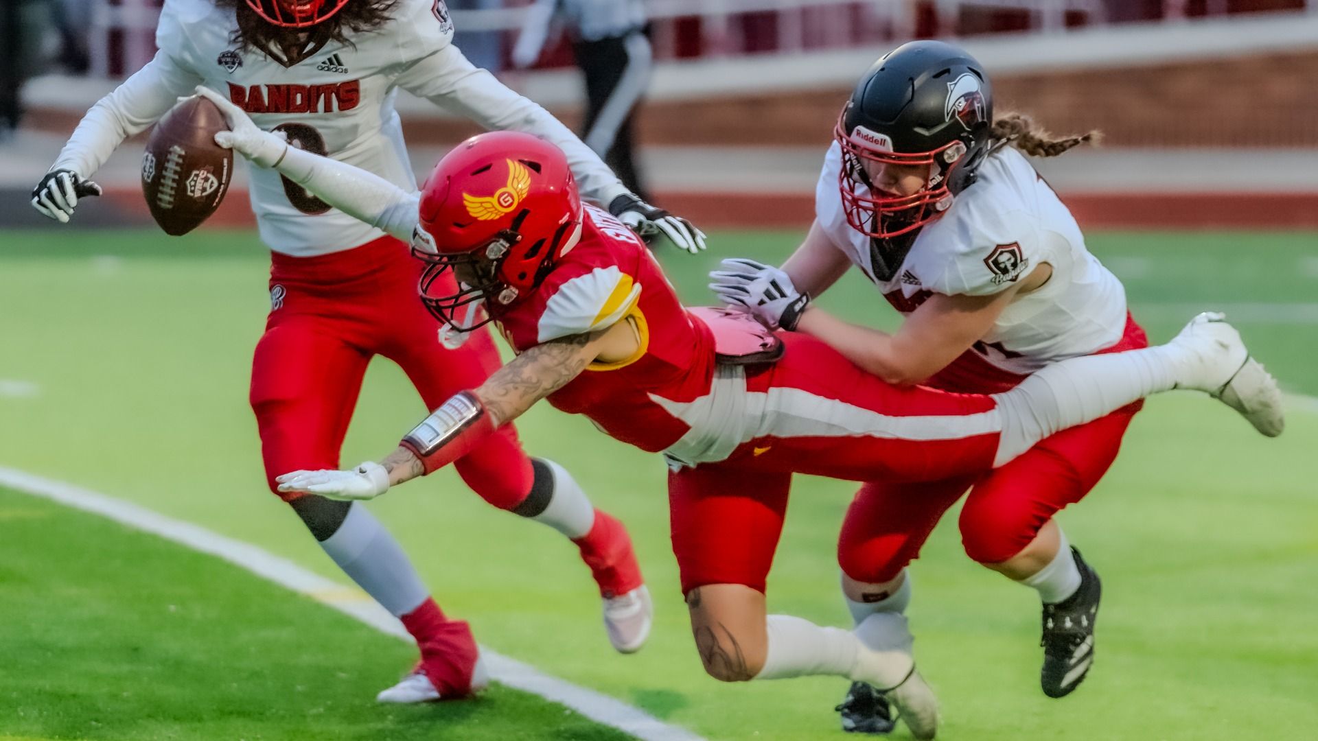 Three football players collide on a green field: a Bandits player in white and red reaches for the football, while a white-uniform defender with a black helmet pushes a Kansas City glory player in red and yellow, who is stretched out with the football over the goal line.