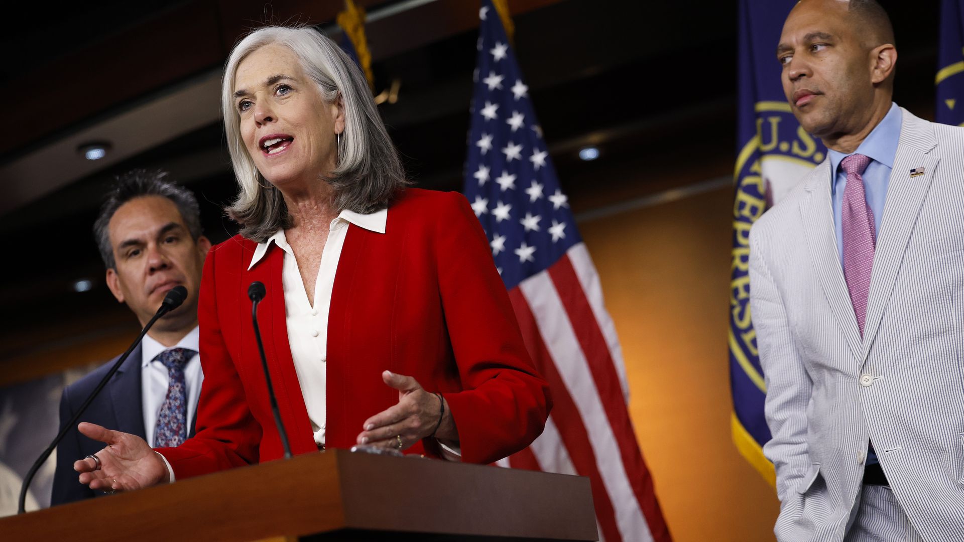 House Minority Whip Katherine Clark, wearing a red blazer, is flanked by Pete Aguilar and Hakeem Jeffries in a TV studio with an American flag in the background.