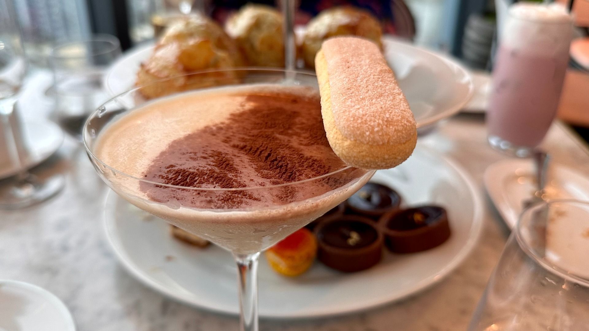 Close-up of a dessert drink in a martini glass topped with cocoa powder and a ladyfinger biscuit, with assorted pastries and a pink drink blurred in the background on a marble table.