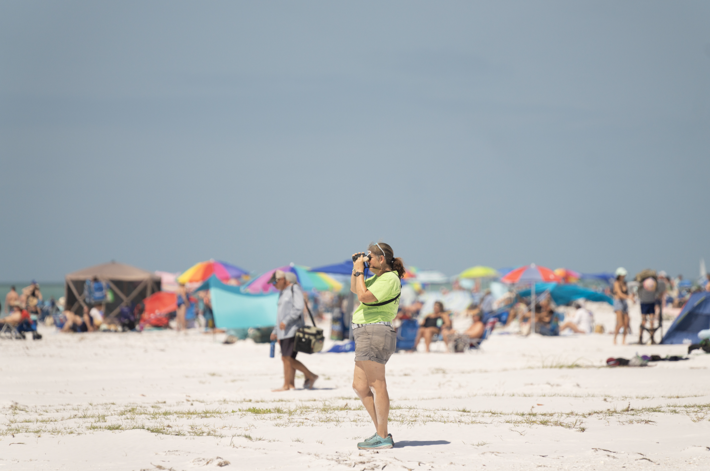 A woman in khaki shorts and a lime-green shirt looking through binoculars at a beach crowded with people and umbrellas.