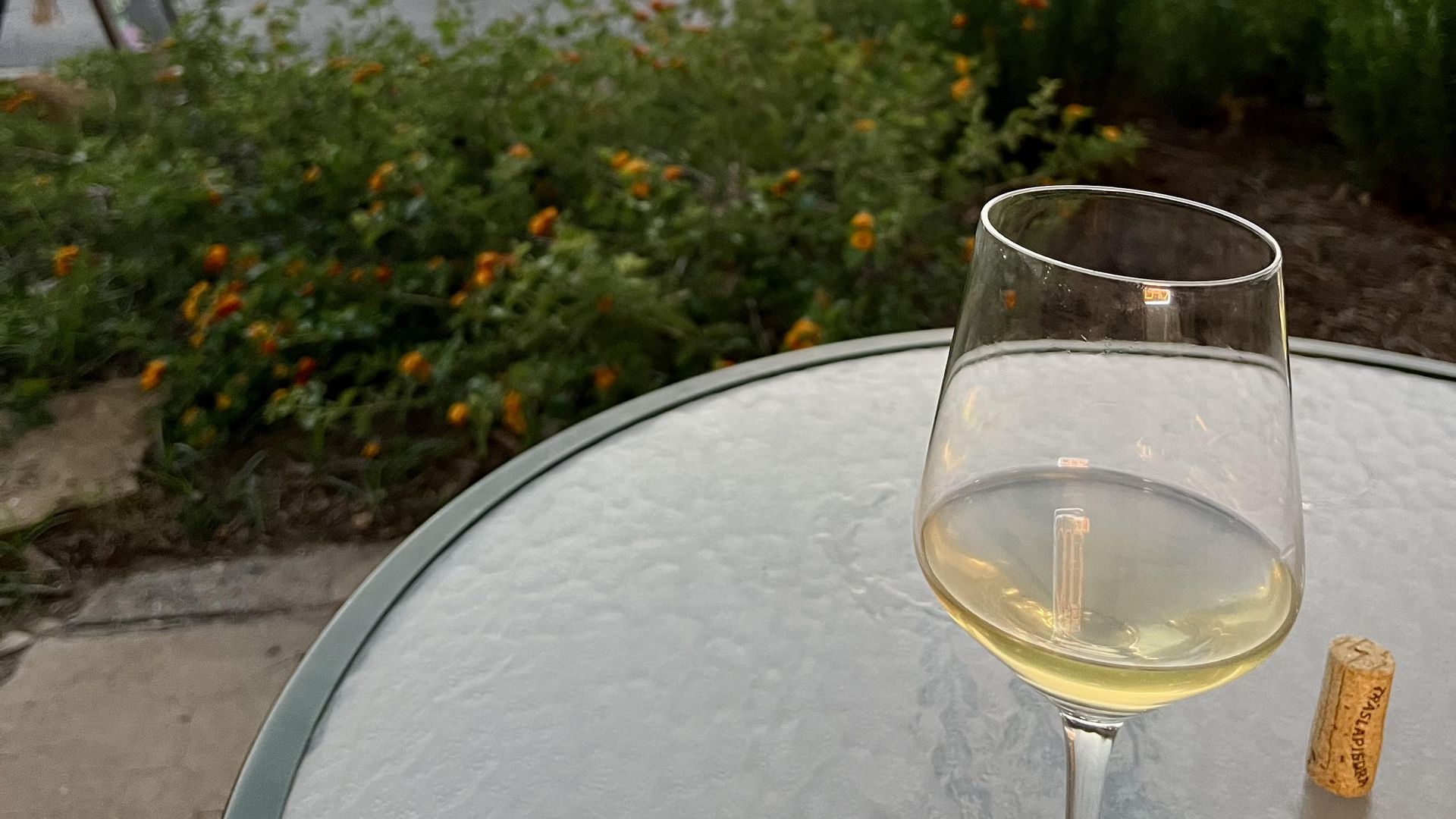 A wine glass with white wine and a cork sits on a glass table outdoors, with a blurred view of a busy store and parked cars in the background at dusk.