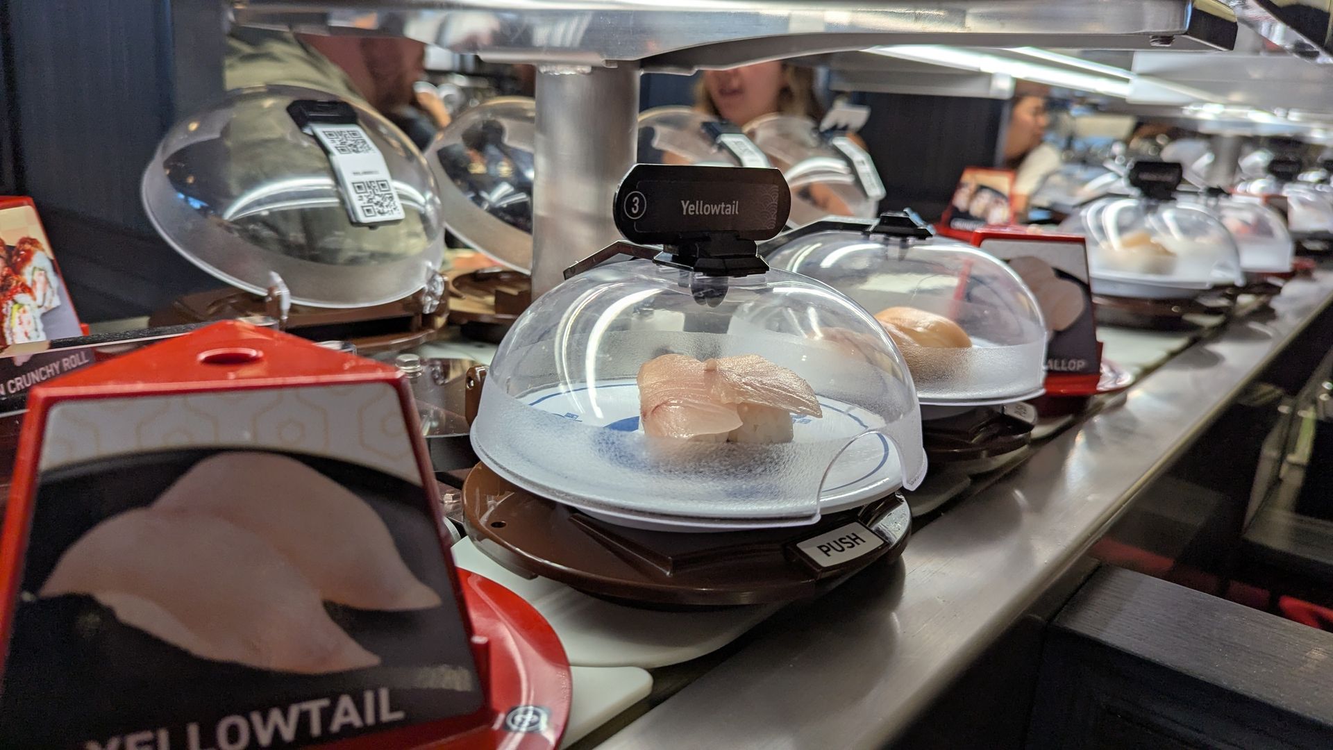 Yellowtail sushi in a clear plastic dome on a conveyer belt.