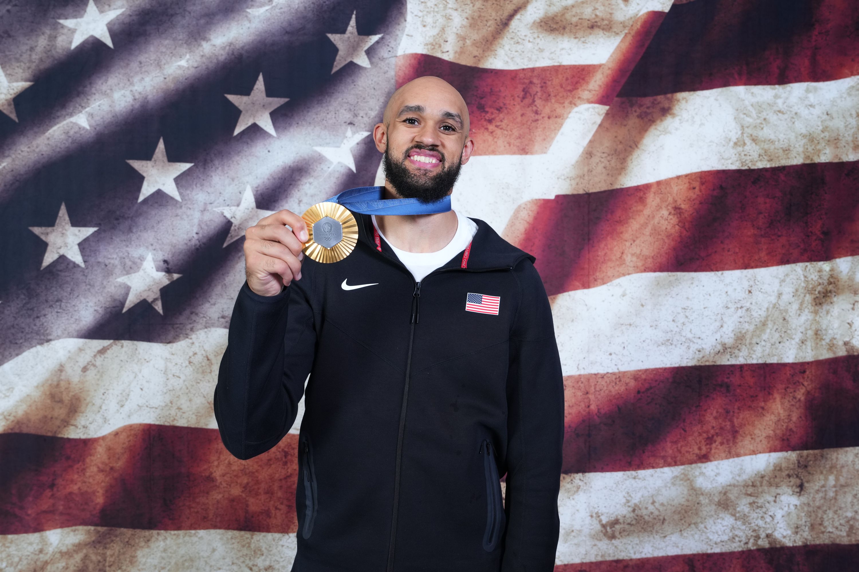 A man holds up a gold medal with an American flag in the background. 
