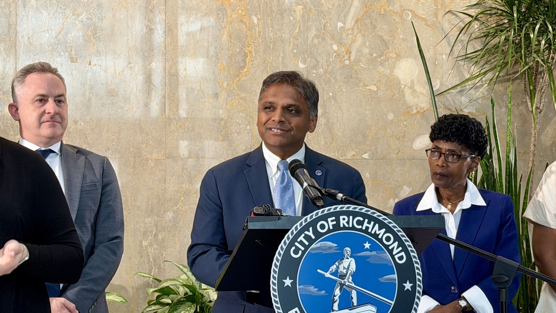 Man in a blue suit speaks at a podium with the City of Richmond seal, flanked by two colleagues, against a marble wall and tall green plants.