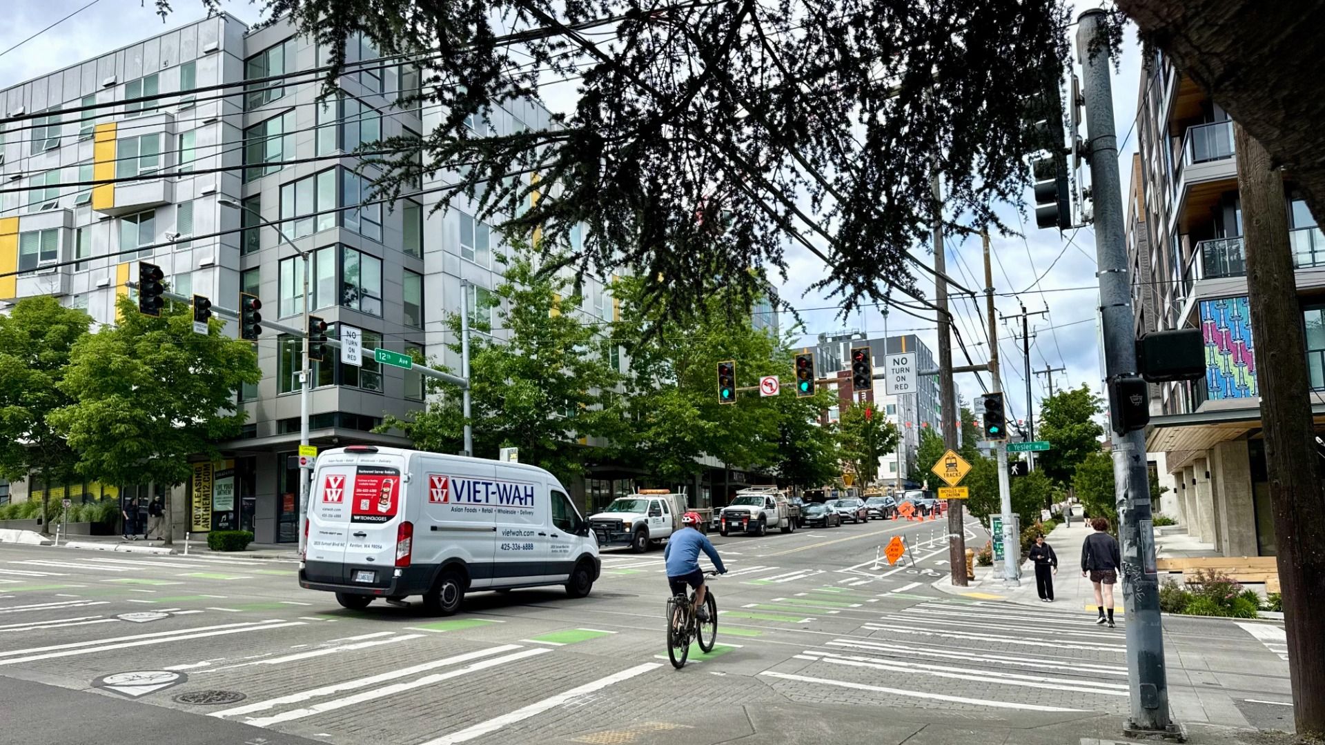 City street intersection with a modern grey apartment building on the left, traffic lights, power lines overhead, a Viet-Wah delivery van, a cyclist in a green bike lane, and pedestrians.