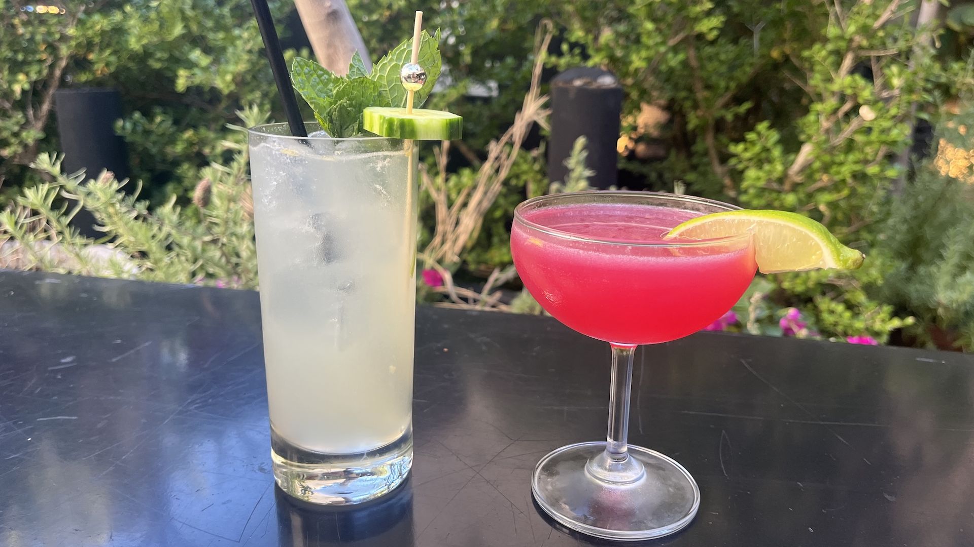 Two cocktails on a black table outdoors: a tall clear drink with ice, mint, cucumber, and black straws, and a vibrant pink drink in a coupe glass garnished with a lime wedge, with greenery background.