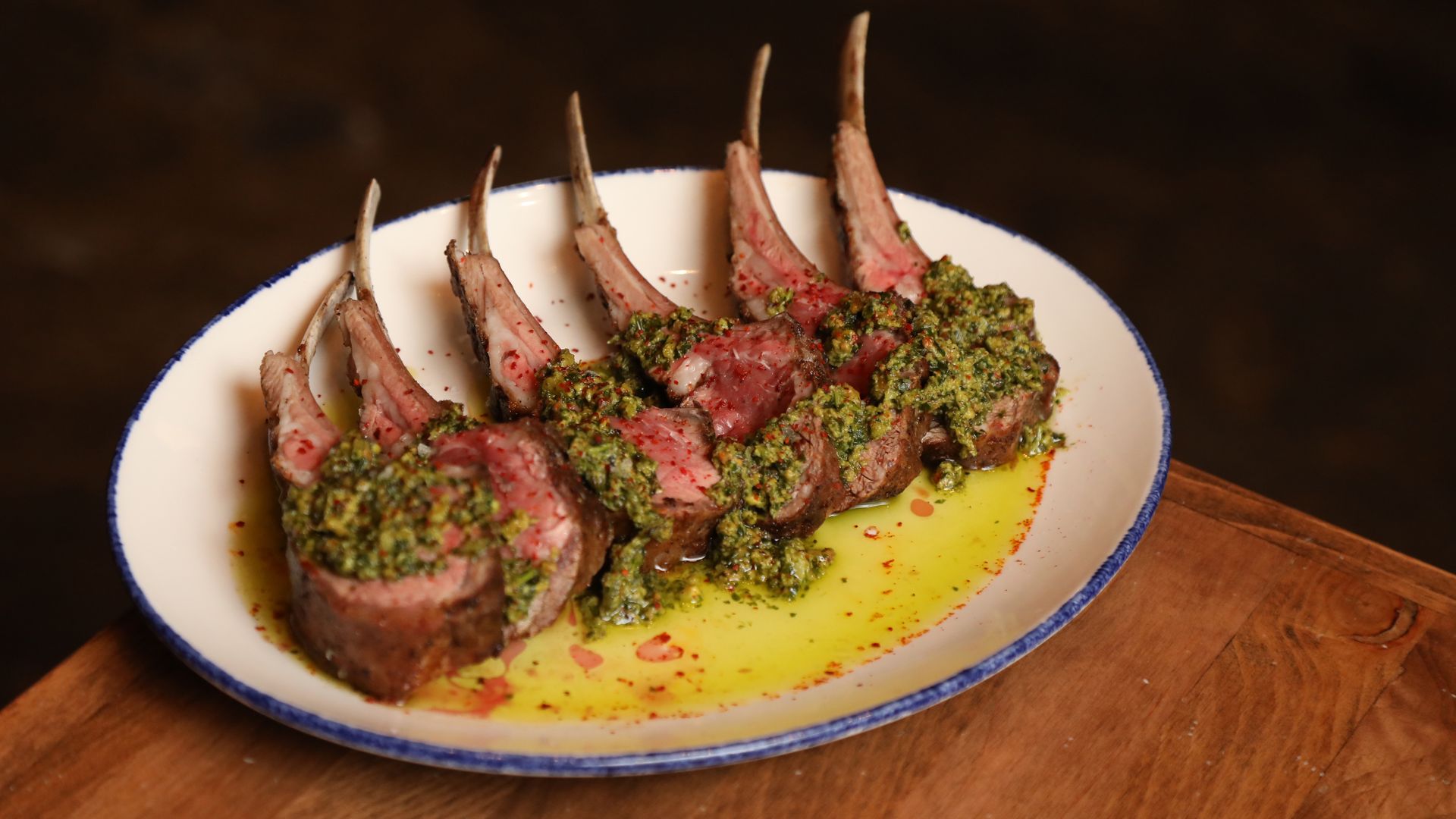 A white plate sits on a wooden table. On the plate is a rare rack of lamb covered with a green sauce. 