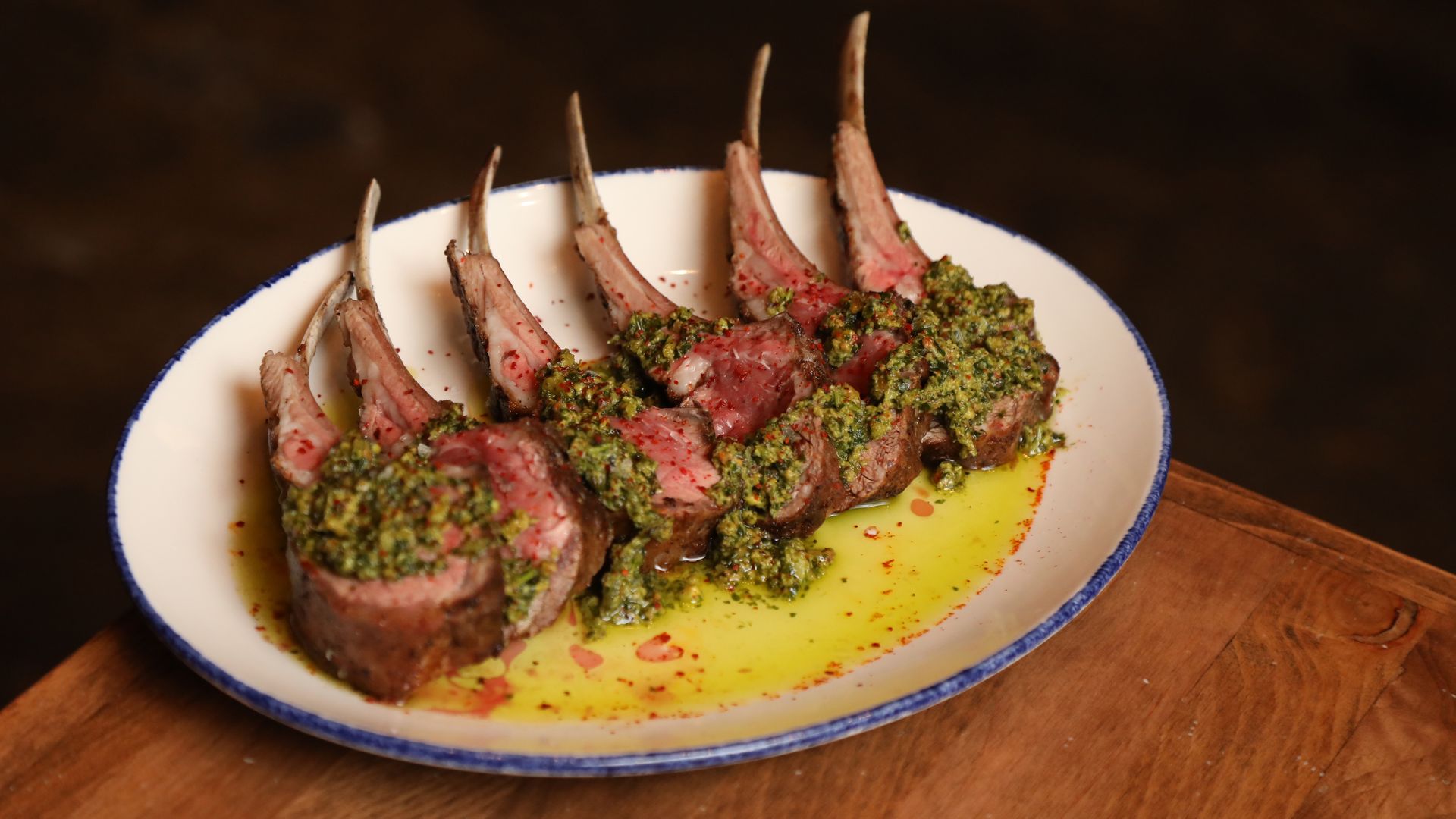 A white plate sits on a wooden table. On the plate is a rare rack of lamb covered with a green sauce.