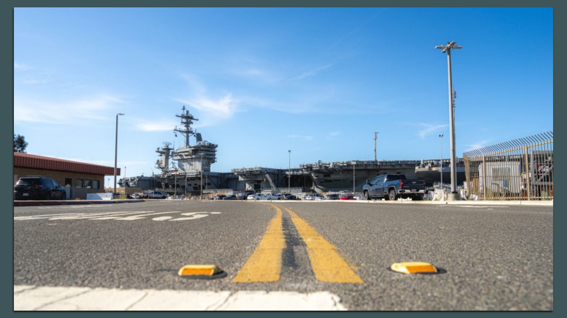 Low-angle view of a large aircraft carrier docked at a pier under a clear blue sky; a road with double yellow lines and parked cars run along the foreground, with a fence and building on the sides.