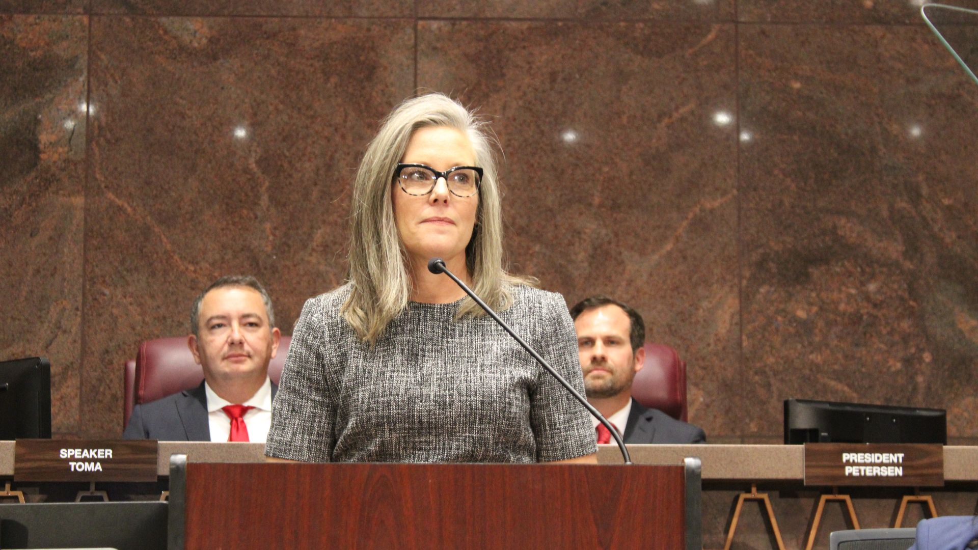 A woman stands at a lectern in front of a microphone with two men sitting behind her. 