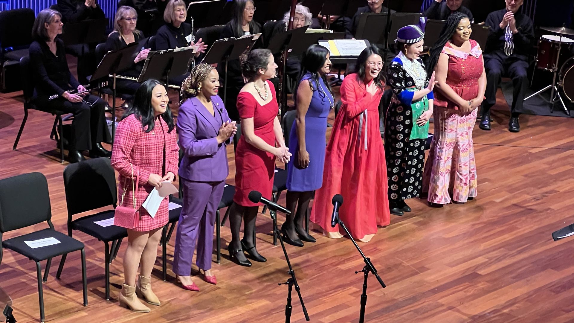 Seven women in colorful dresses and business wear stand on a stage and acknowledge applause