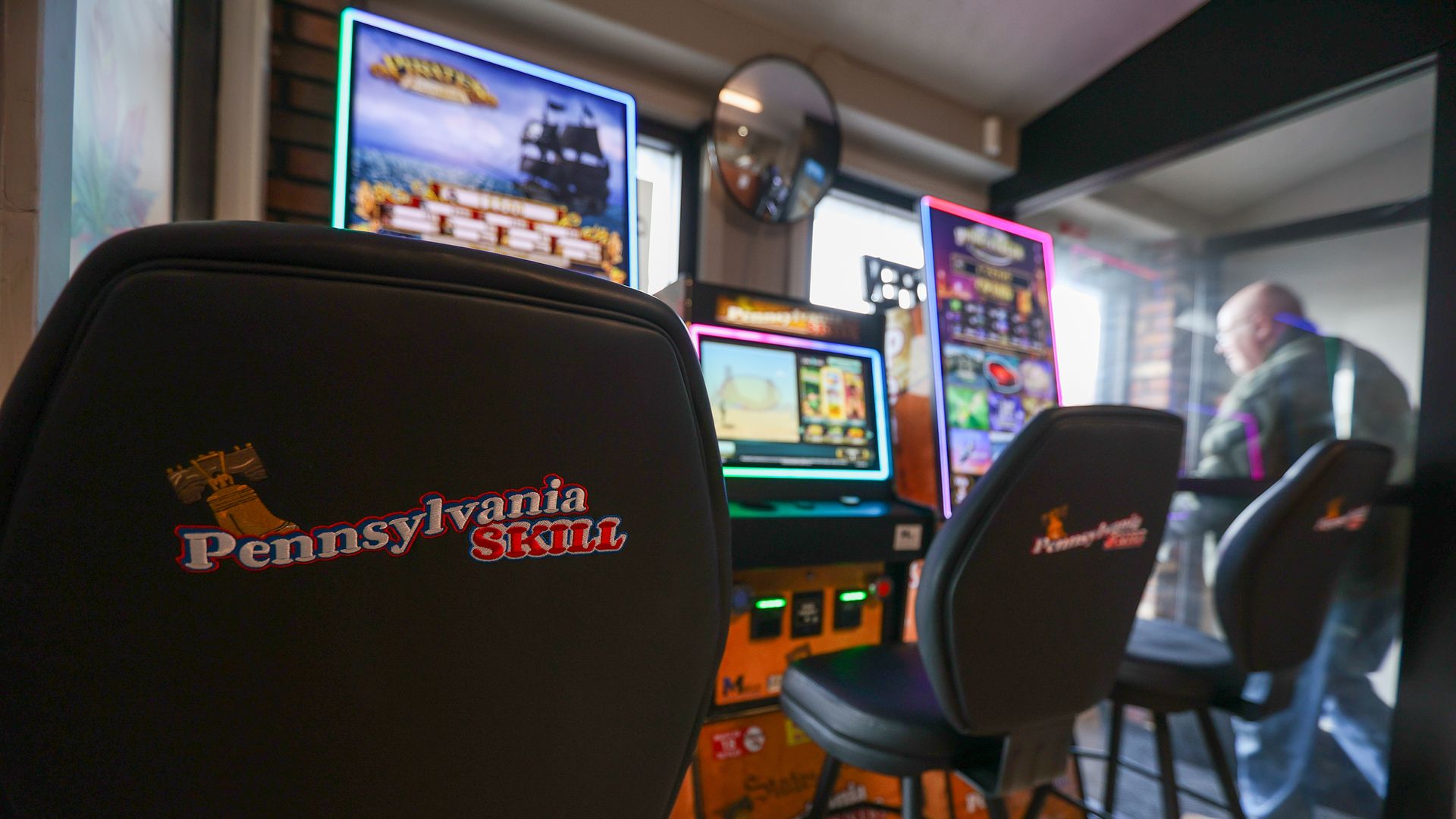 Indoor skill games area with multiple illuminated gaming monitors in bright blue and pink neon frames. Foreground chairs read "Pennsylvania Skill". A man in a hoodie stands by a glass wall on the right.